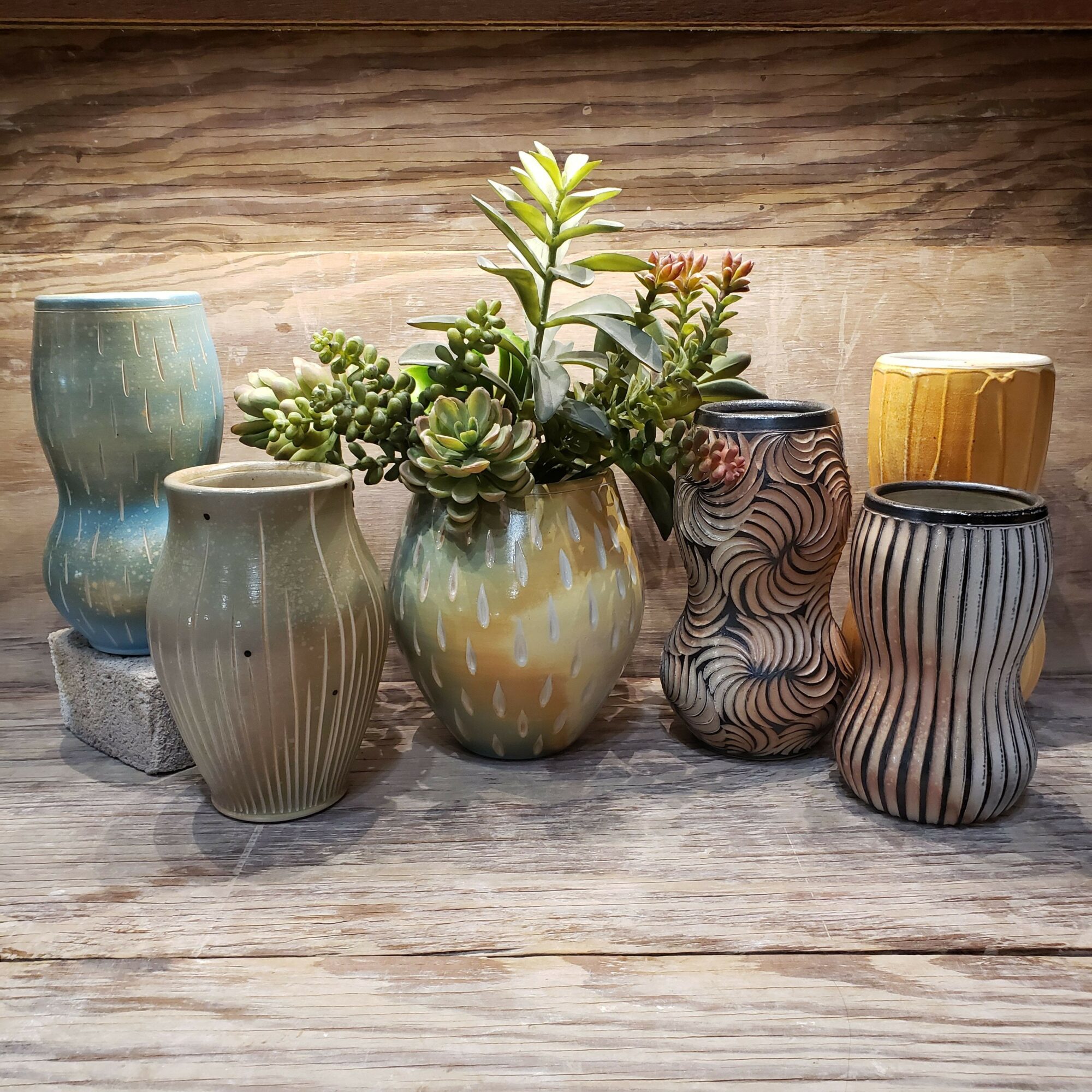 Six decorative vases with plants on a wooden surface, various shapes and patterns, some with greenery, arranged vertically.