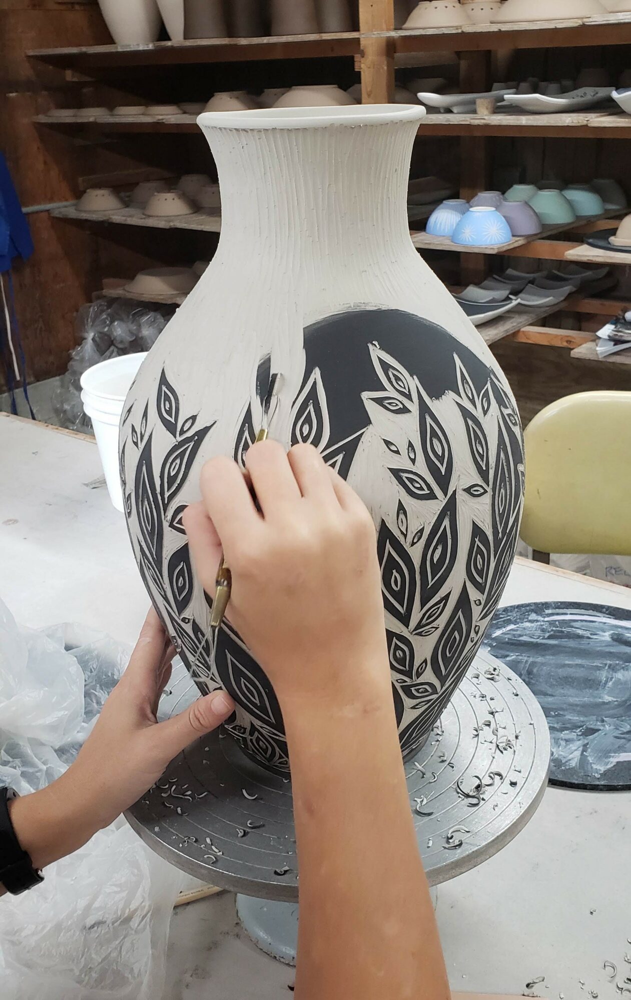 Person decorating a large ceramic vase with black leaf patterns in a workshop.