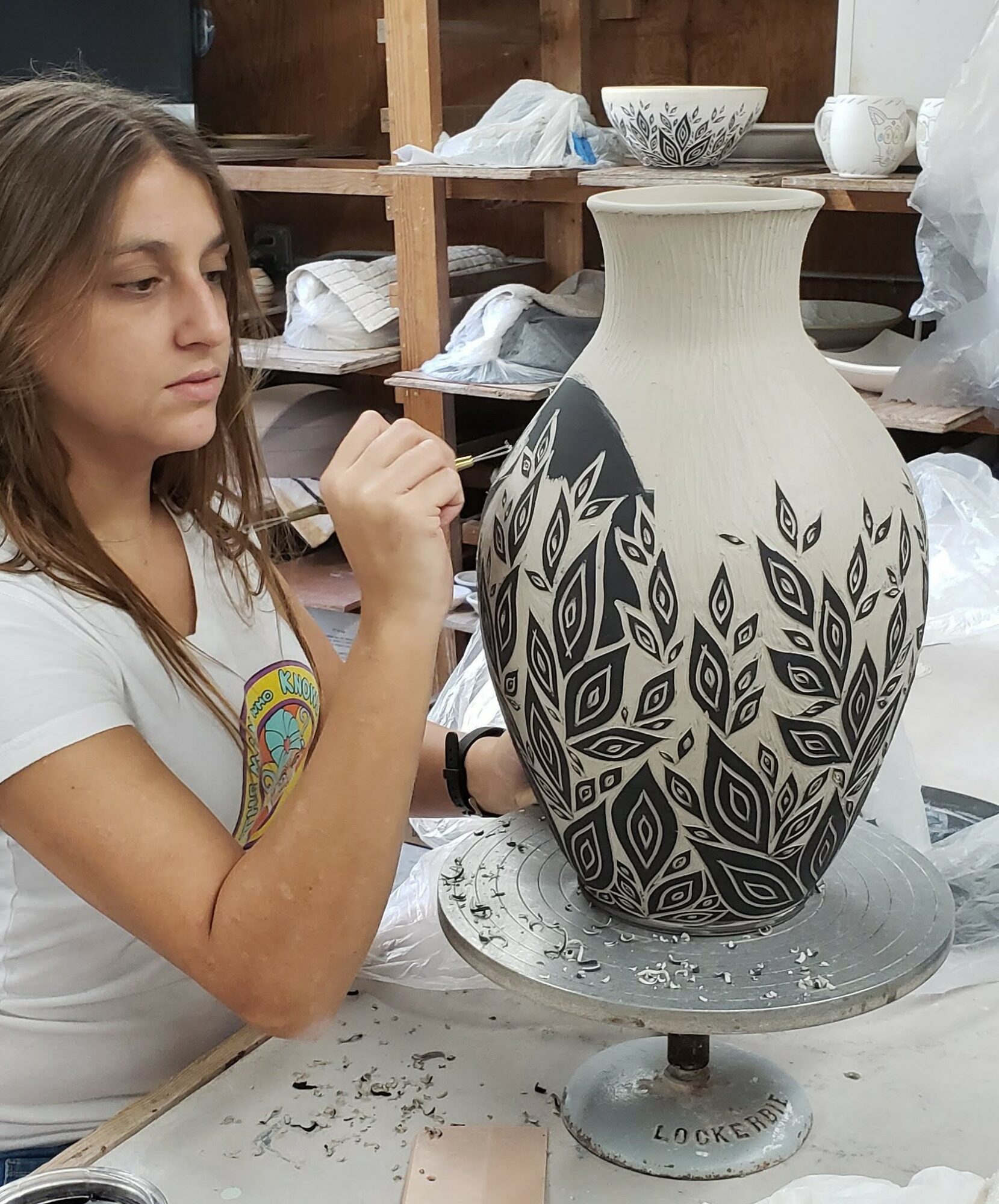 Young girl painting a large black and white patterned vase in a workshop, with shelves and supplies in the background.