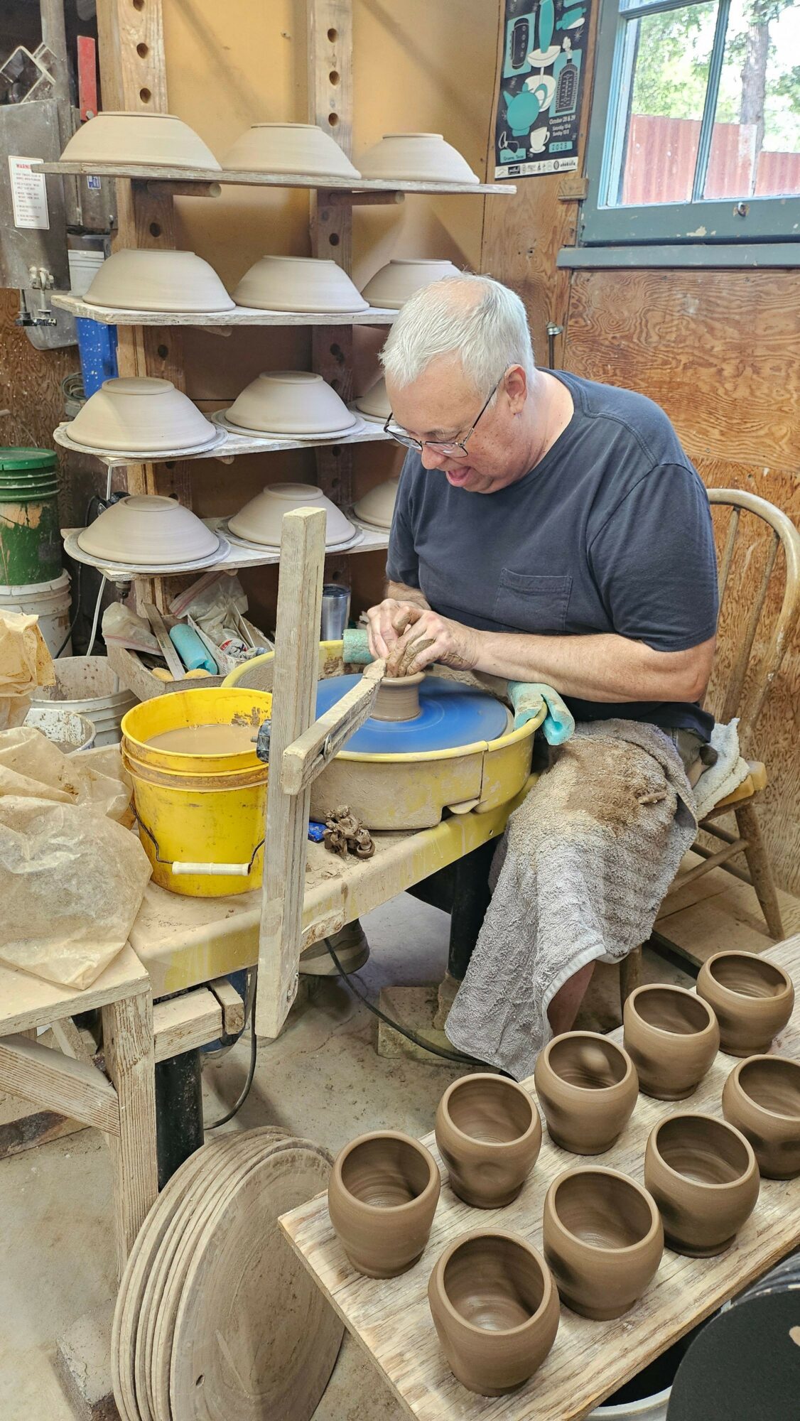 Person working with pottery tools in a workshop, surrounded by clay pots and pottery supplies.