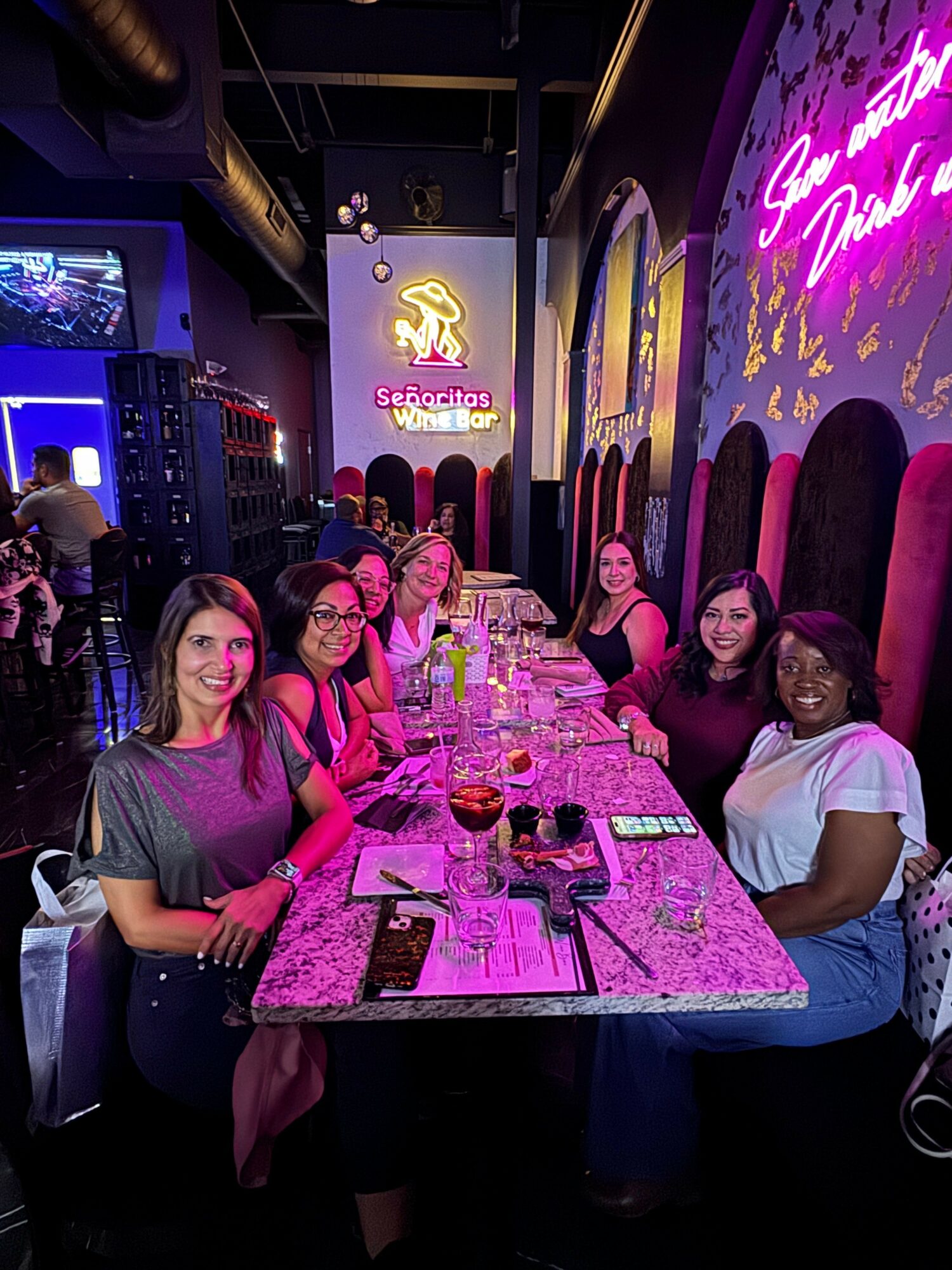 Group of people sitting at a table in a dimly lit restaurant or bar, with a neon sign on the wall behind them.