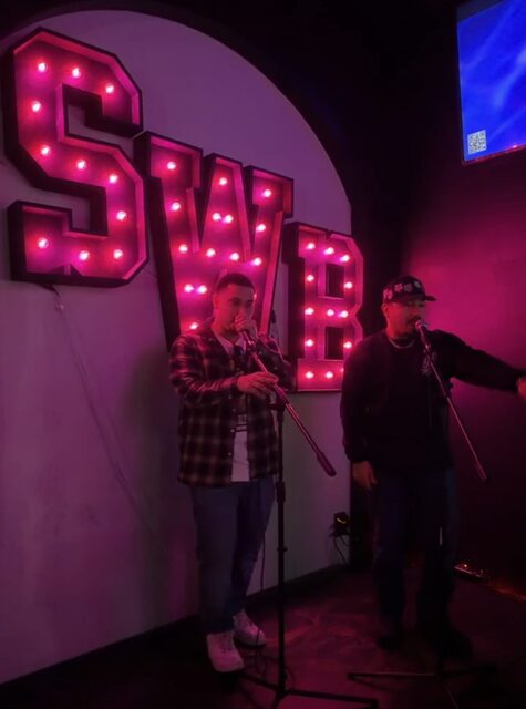 Two people stand on stage with microphones, in front of a large illuminated sign spelling 'SWIR' with pink lights.