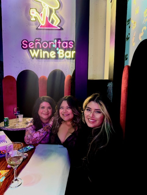 Three women sitting at a table in a wine bar, smiling, with a neon sign that reads 'Señoritas Wine Bar' behind them.