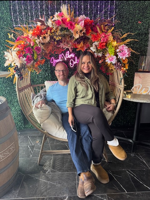 Two people sitting on a circular swing decorated with colorful flowers, with a neon sign behind them, in an indoor setting.