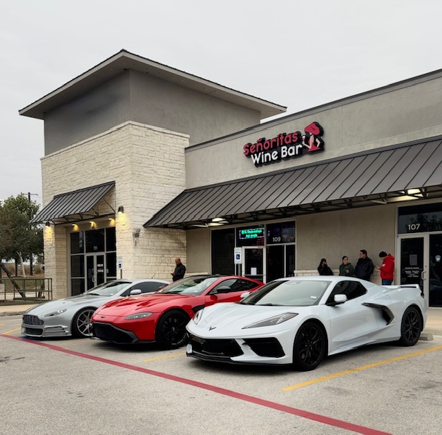 Three sports cars parked outside a wine bar, with people near the entrance, in front of a shopping center.