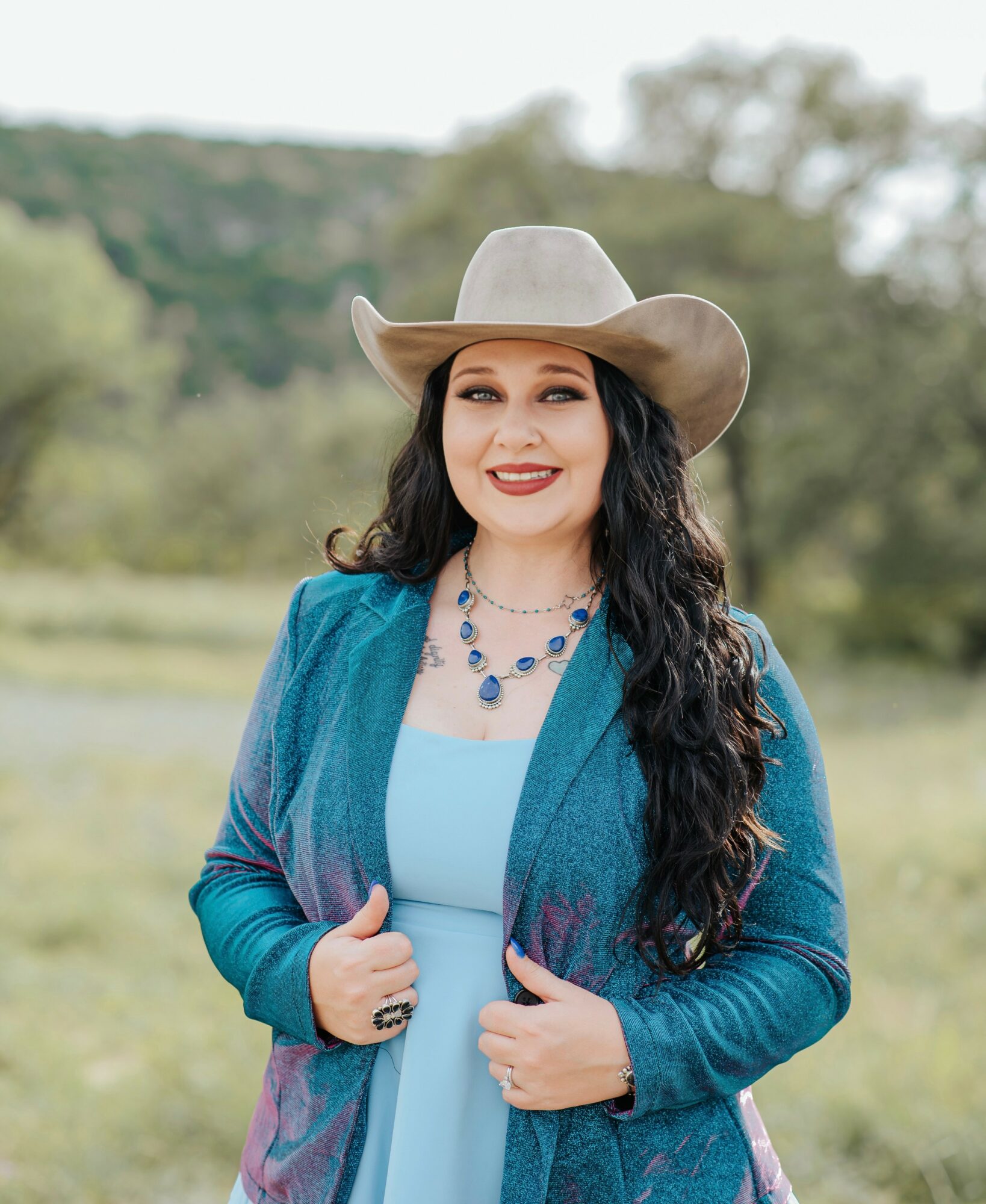 Woman in a cowboy hat and blue outfit outdoors, smiling, with trees and sky in background.