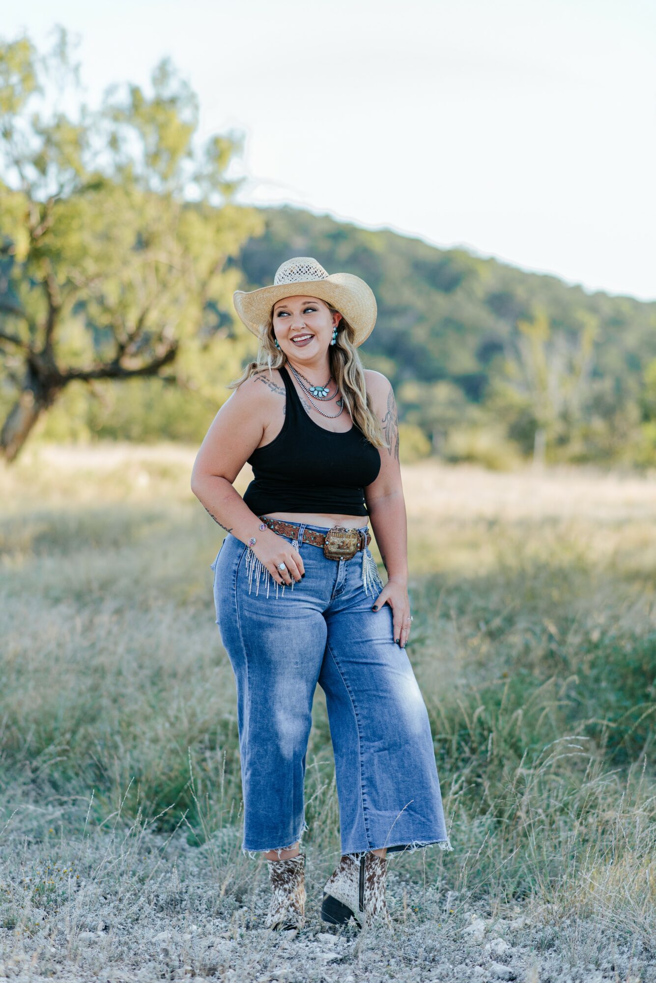 Woman standing outdoors in a grassy field, wearing a wide-brimmed hat, black top, and jeans, smiling.