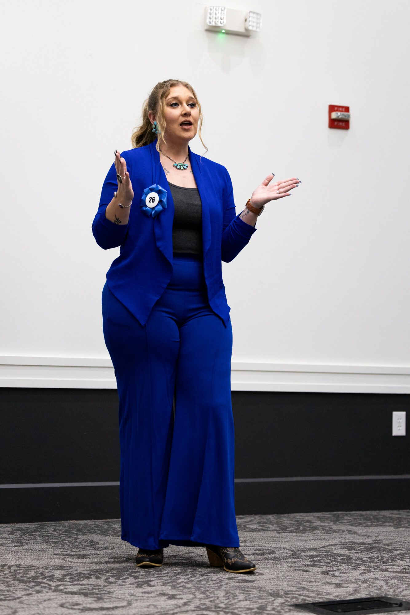 Woman in a blue suit speaking with hands raised, standing against a white wall, carpeted floor, and fire alarm.