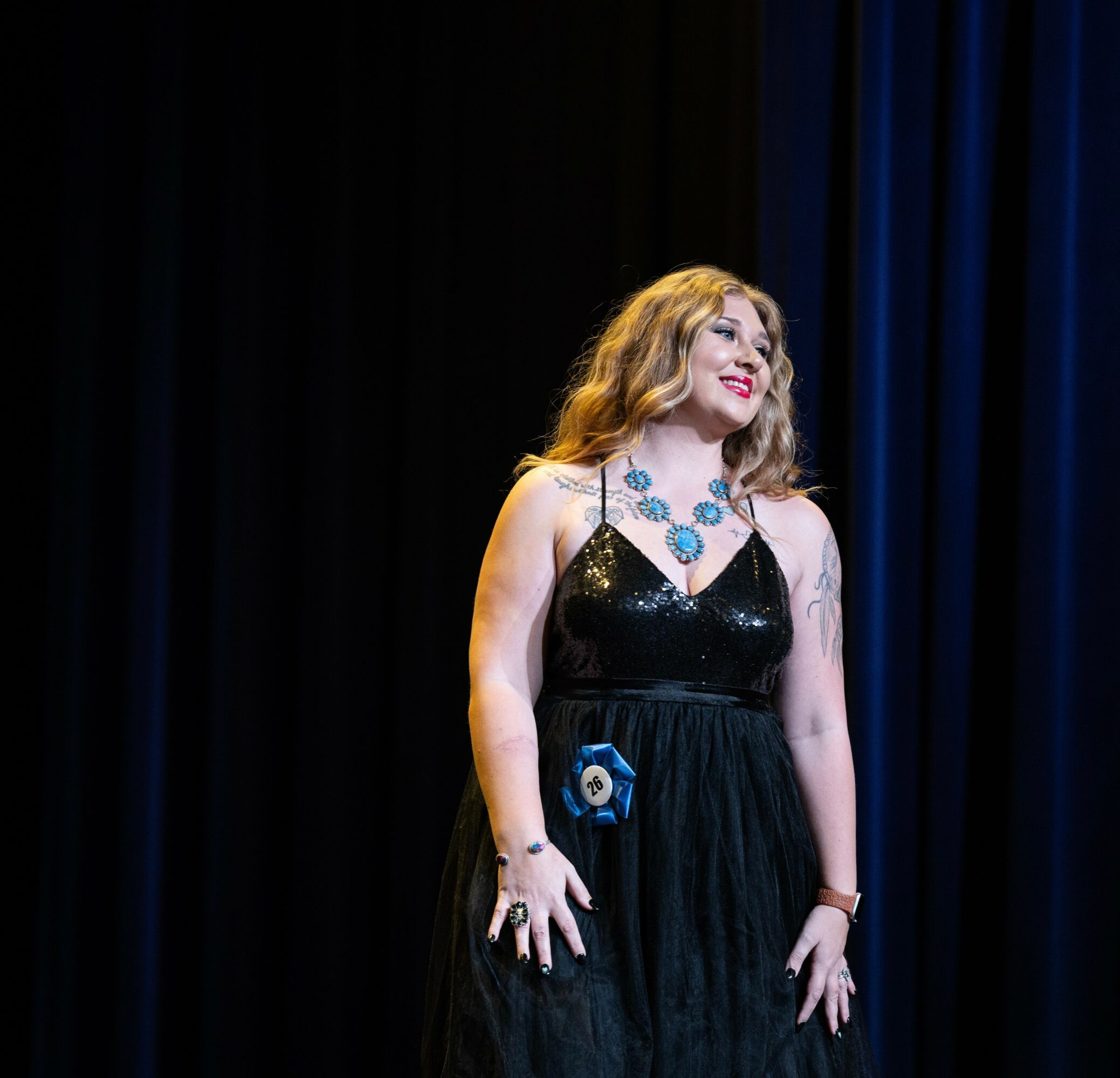 Woman with wavy blonde hair in a black dress and blue jewelry stands on stage with dark curtains behind her.