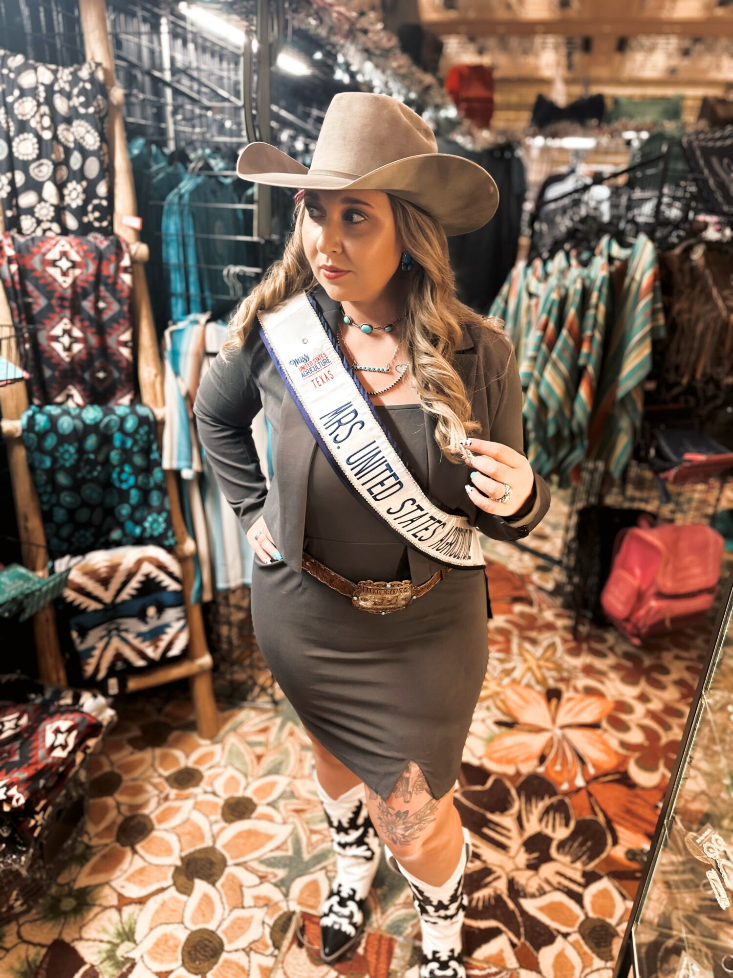 Woman in a hat and sash shopping in a store with clothing and accessories, standing on patterned floor.