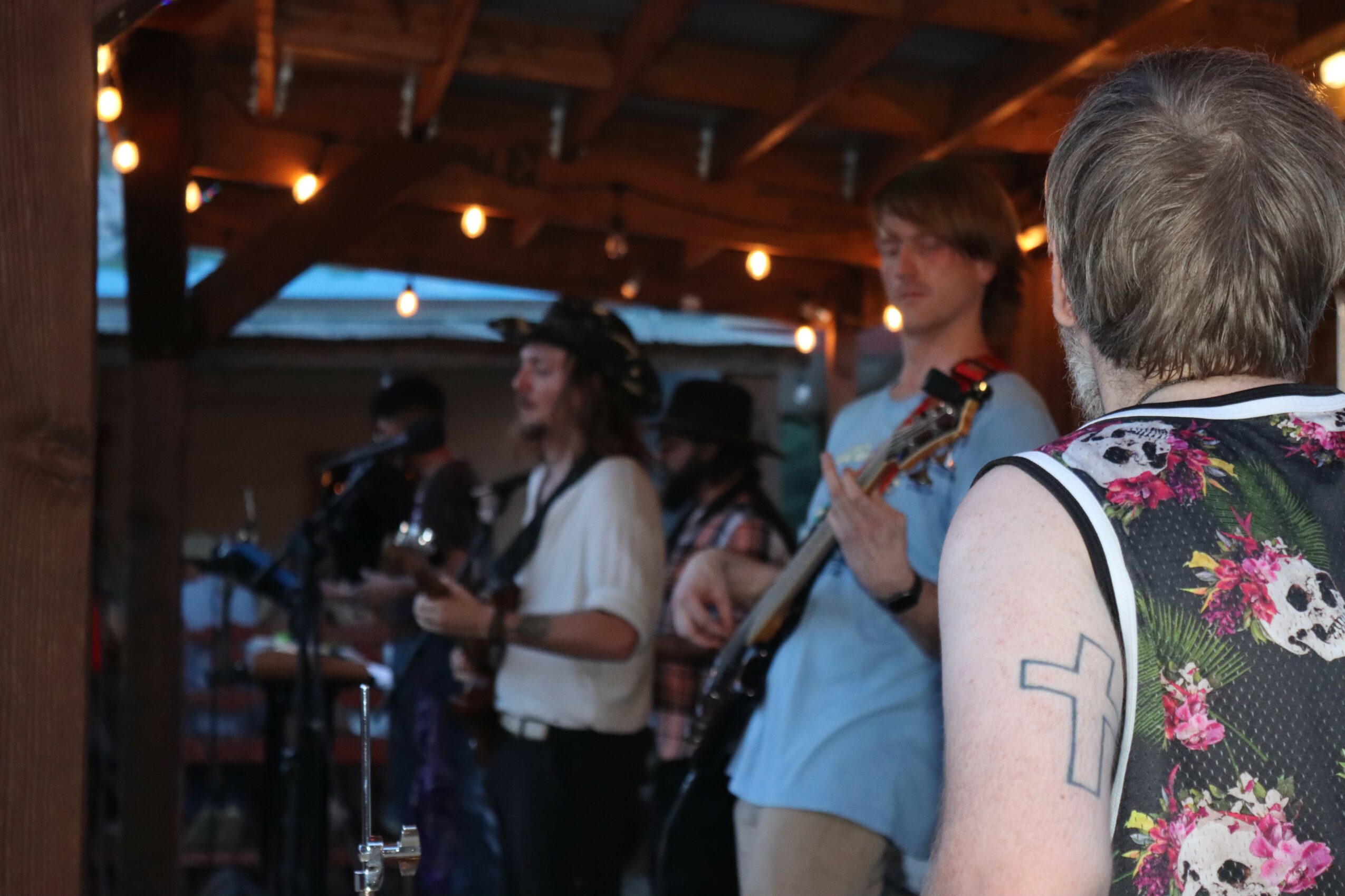 Group of people playing musical instruments in a wooden outdoor setting during evening, with string lights overhead.
