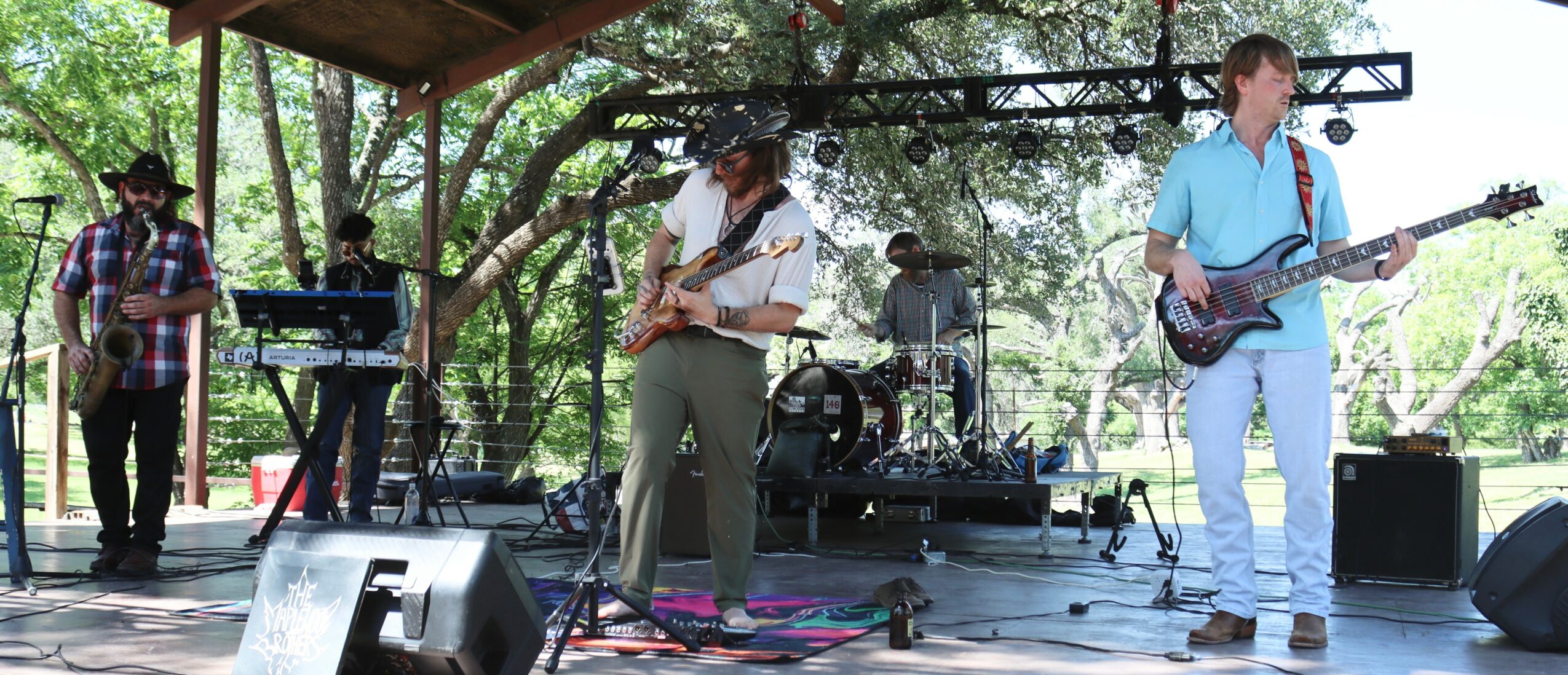 Band performing outdoors on stage with trees in background, four musicians playing instruments, including guitar and keyboard.