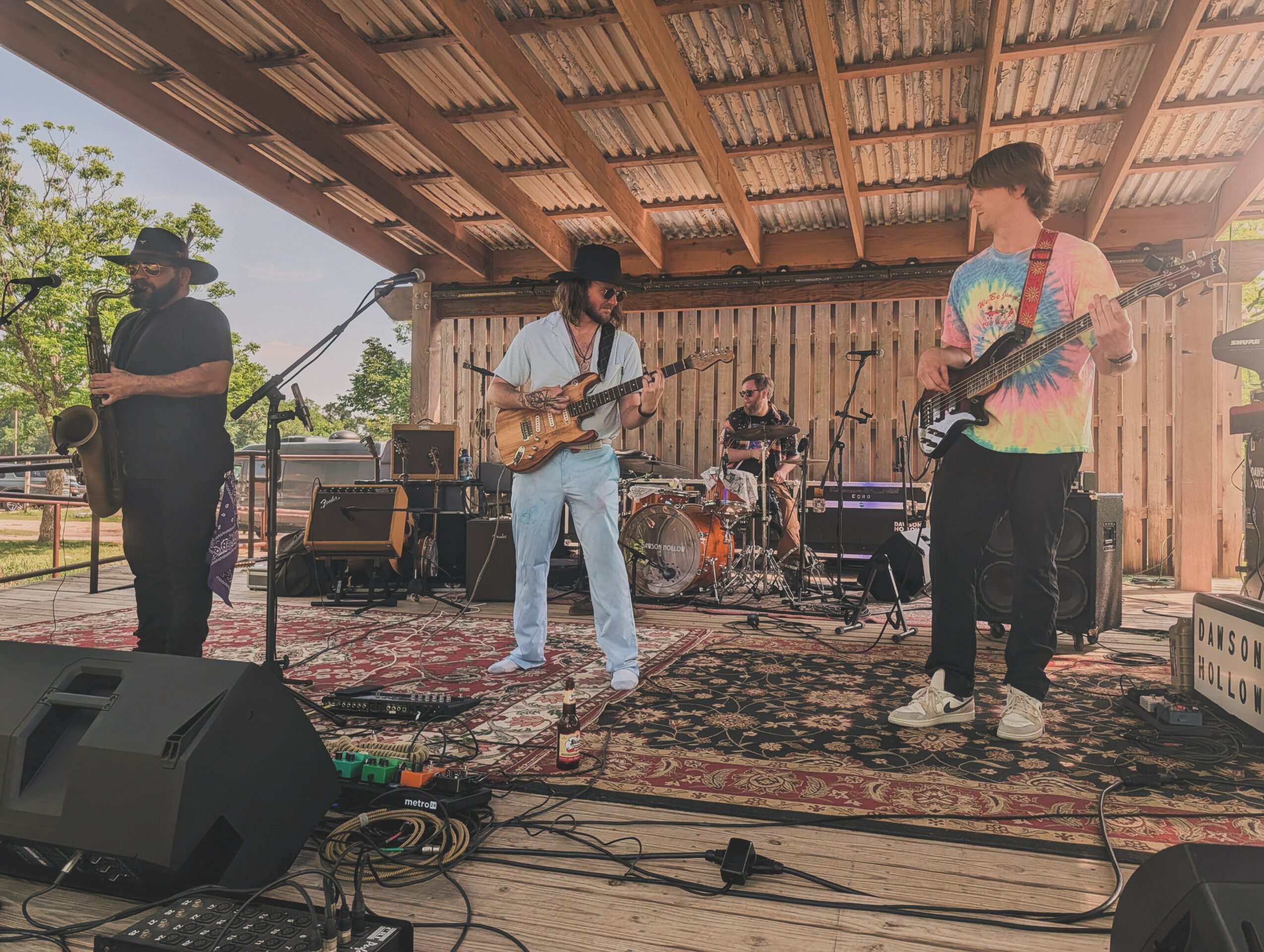 Band performing on stage with four musicians playing guitars and drums, under a wooden roof, outdoor setting, sound equipment visible.