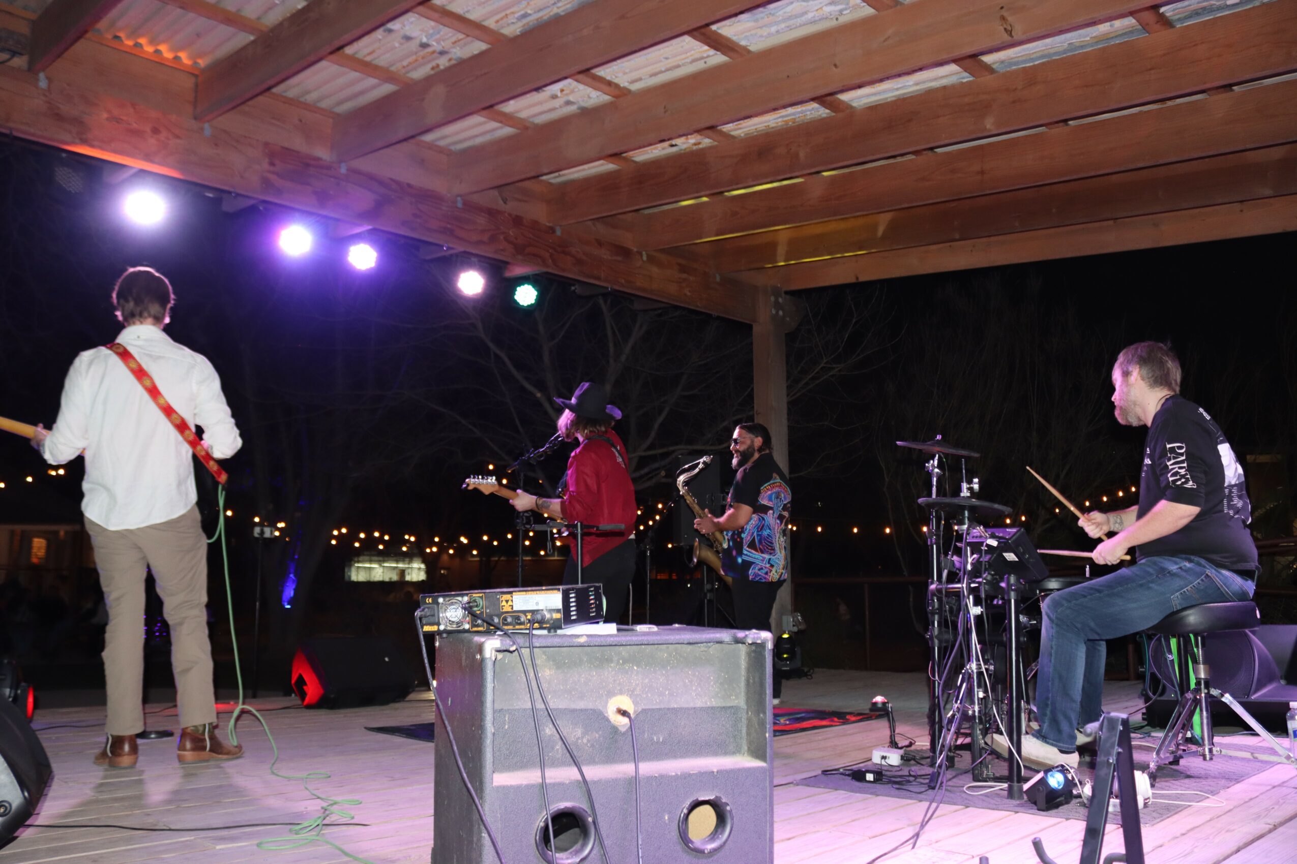 Four musicians perform on a stage under a wooden roof at night, with city lights in the background.