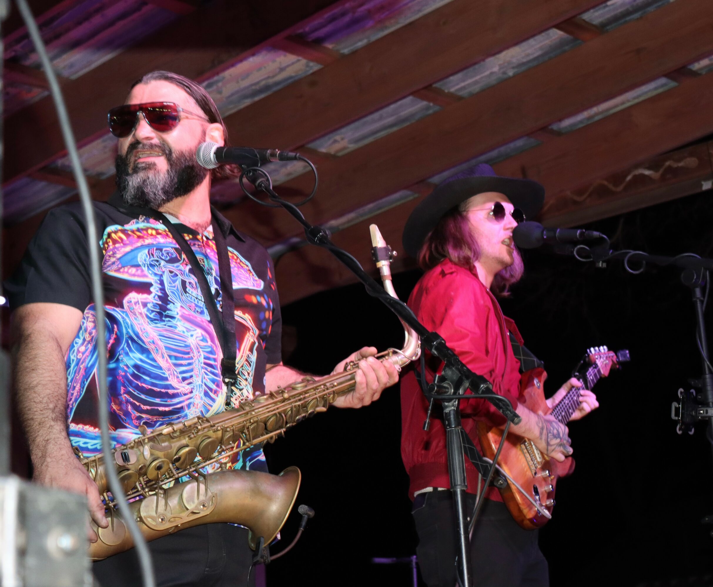Two musicians perform on stage, one playing saxophone and the other guitar, both wearing sunglasses and colorful clothing.