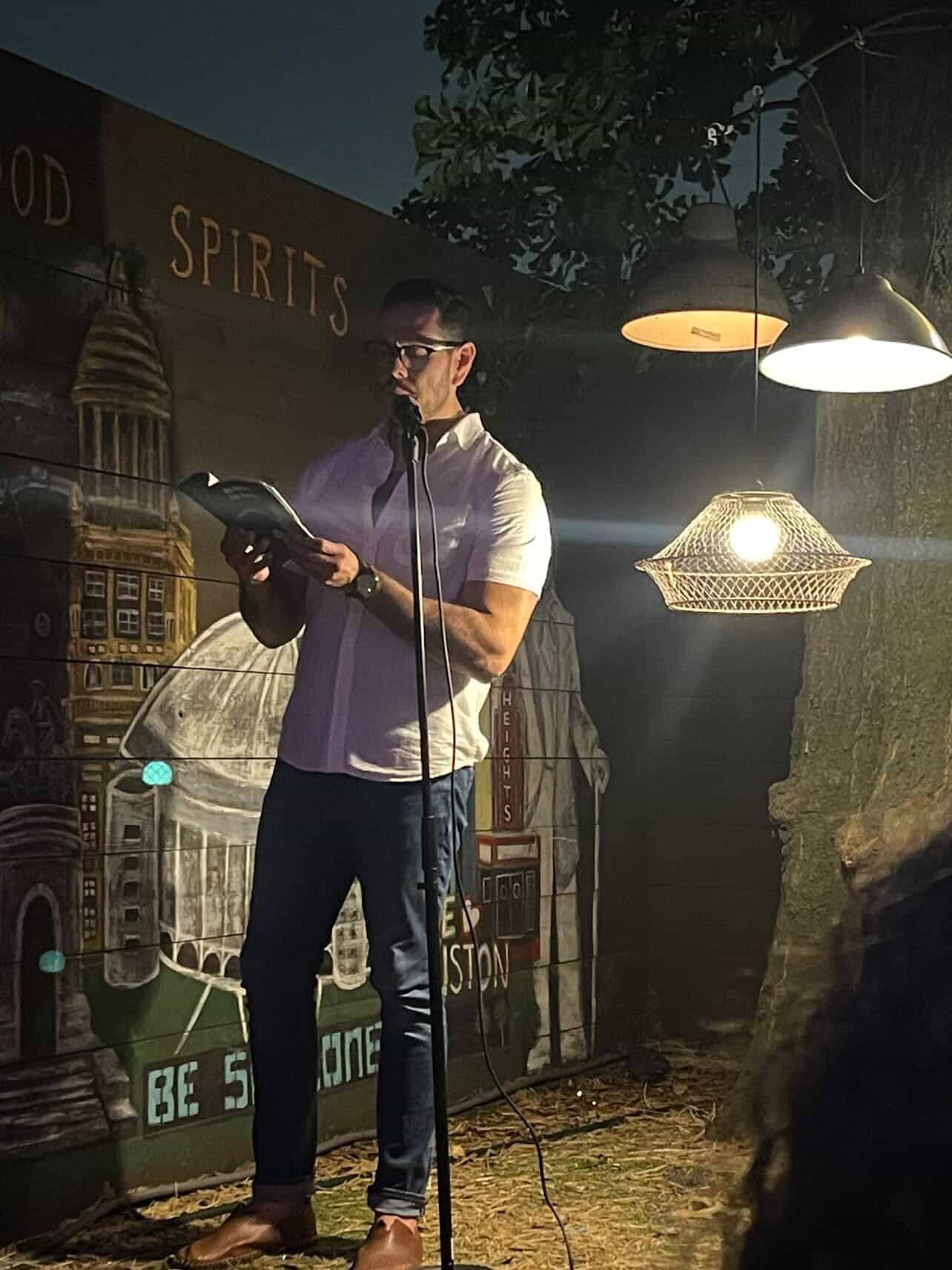 Man wearing glasses and headphones reads a book in a dimly lit room with hanging lights and a mural background.