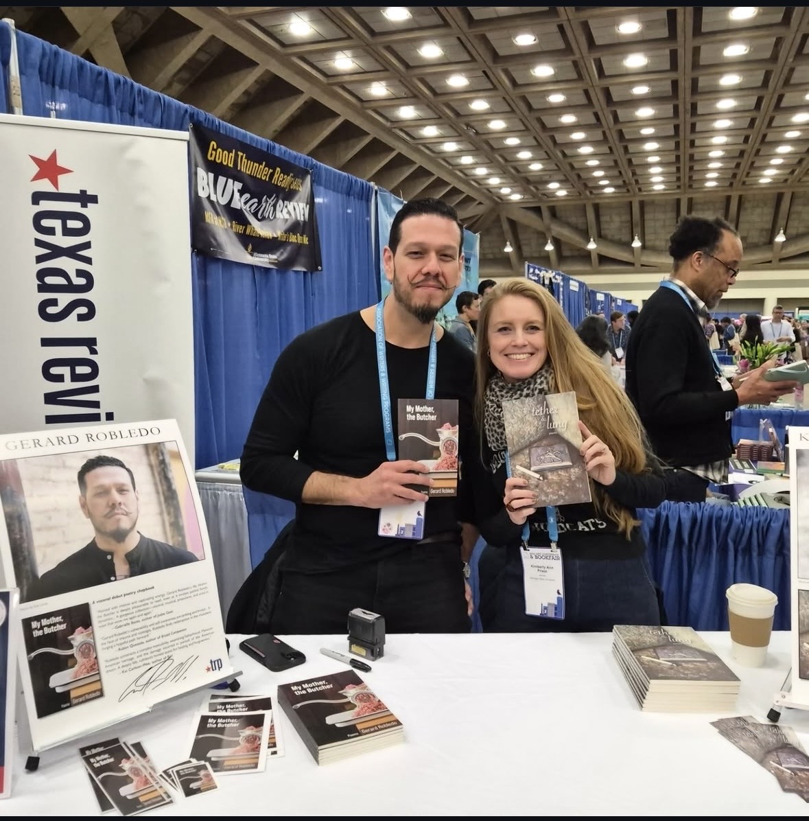 Two people smiling and holding books at a booth in a large indoor event space, with tables and banners in the background.