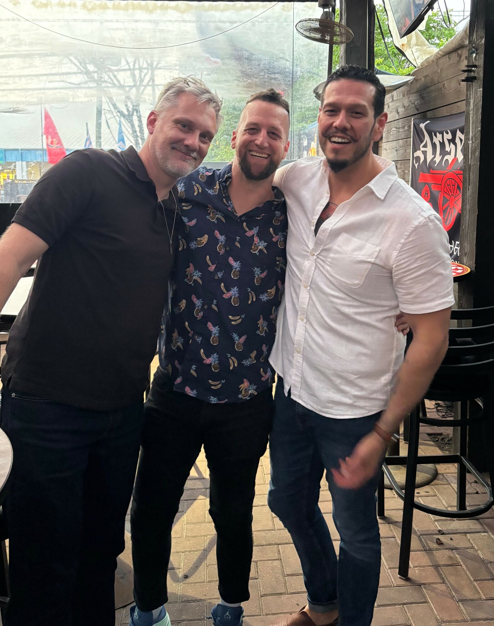 Three men standing together outdoors, smiling, with a tent and chairs in the background.
