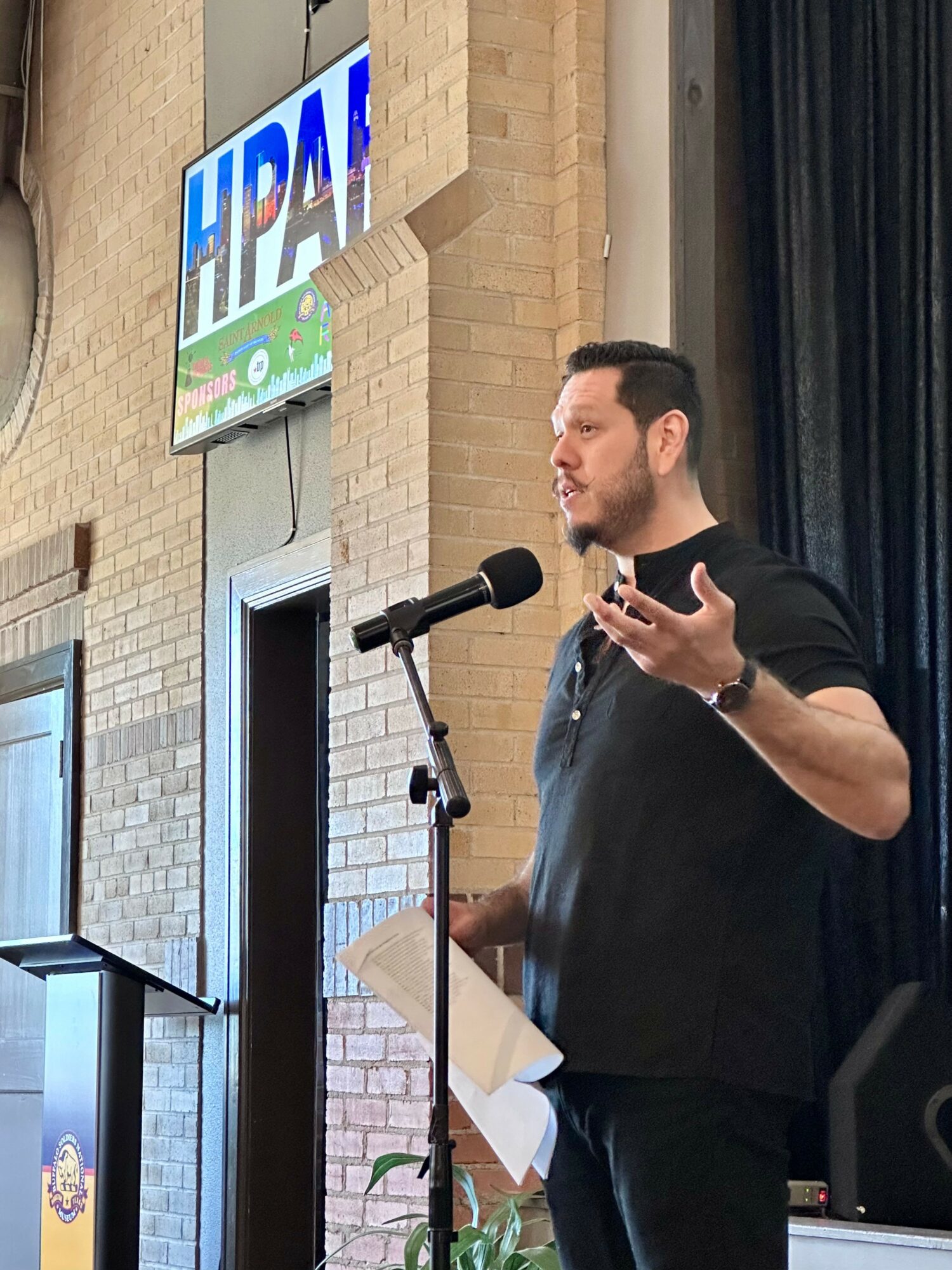 Man speaking at a microphone during a presentation or speech, holding papers, with a brick wall and a screen in the background.