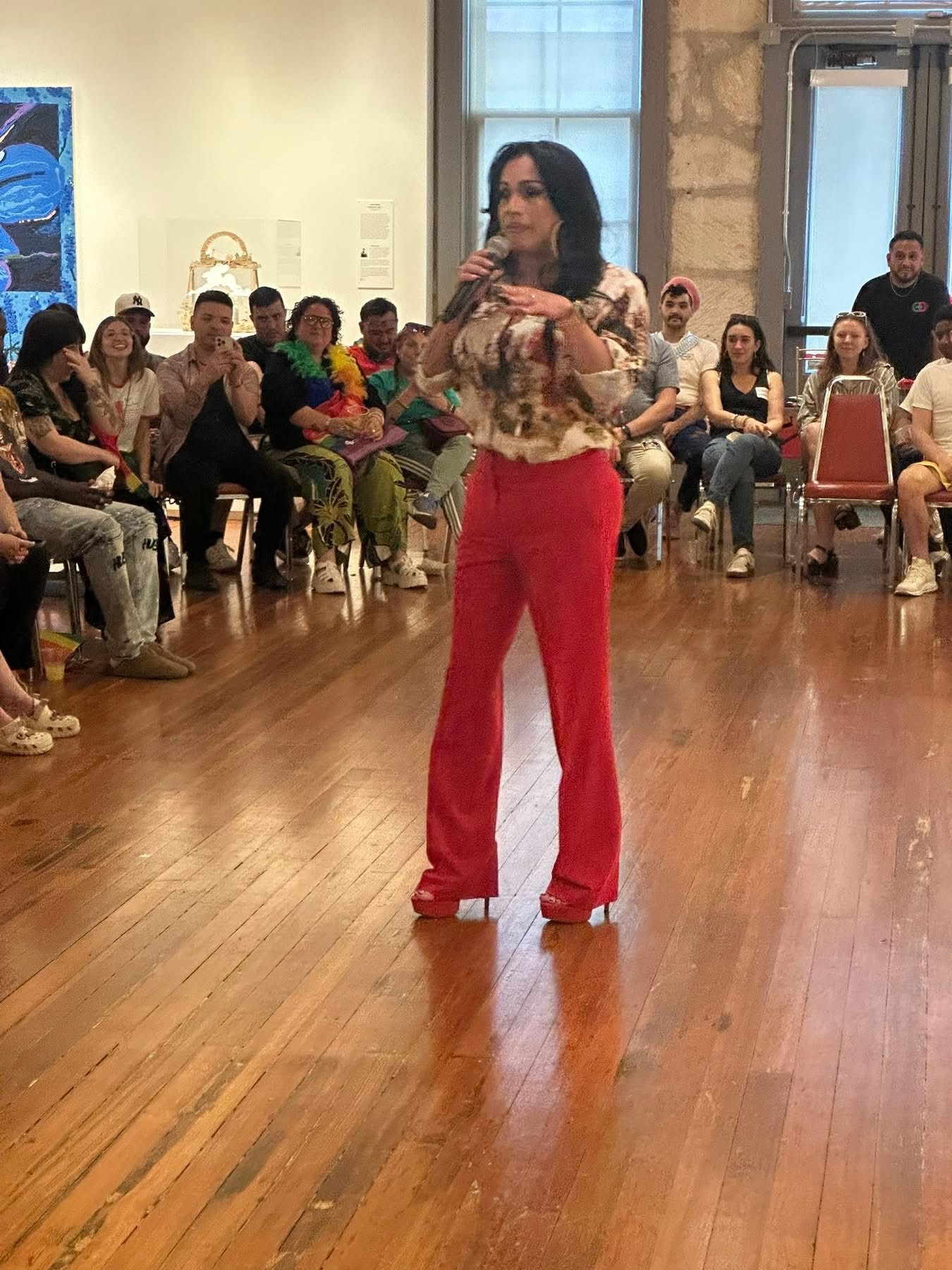 Woman standing in front of seated audience, speaking into microphone, in a room with wooden floor and large window.