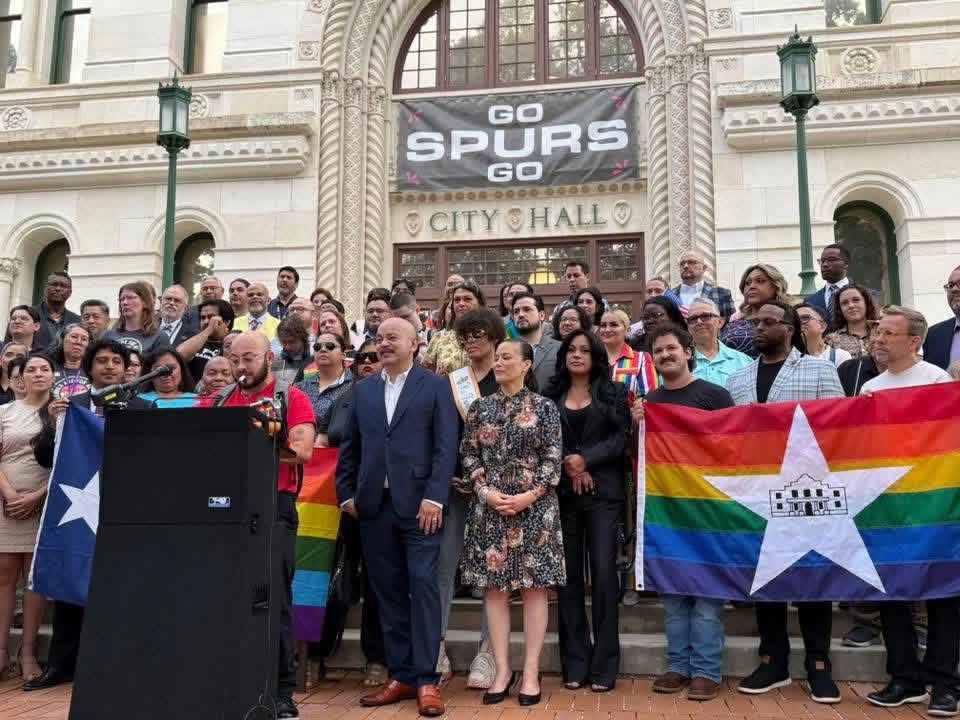 Group of people gathered outside a building holding a rainbow flag with a star and a Puerto Rican flag, with a large screen displaying 'GO SPURS GO' in the background.