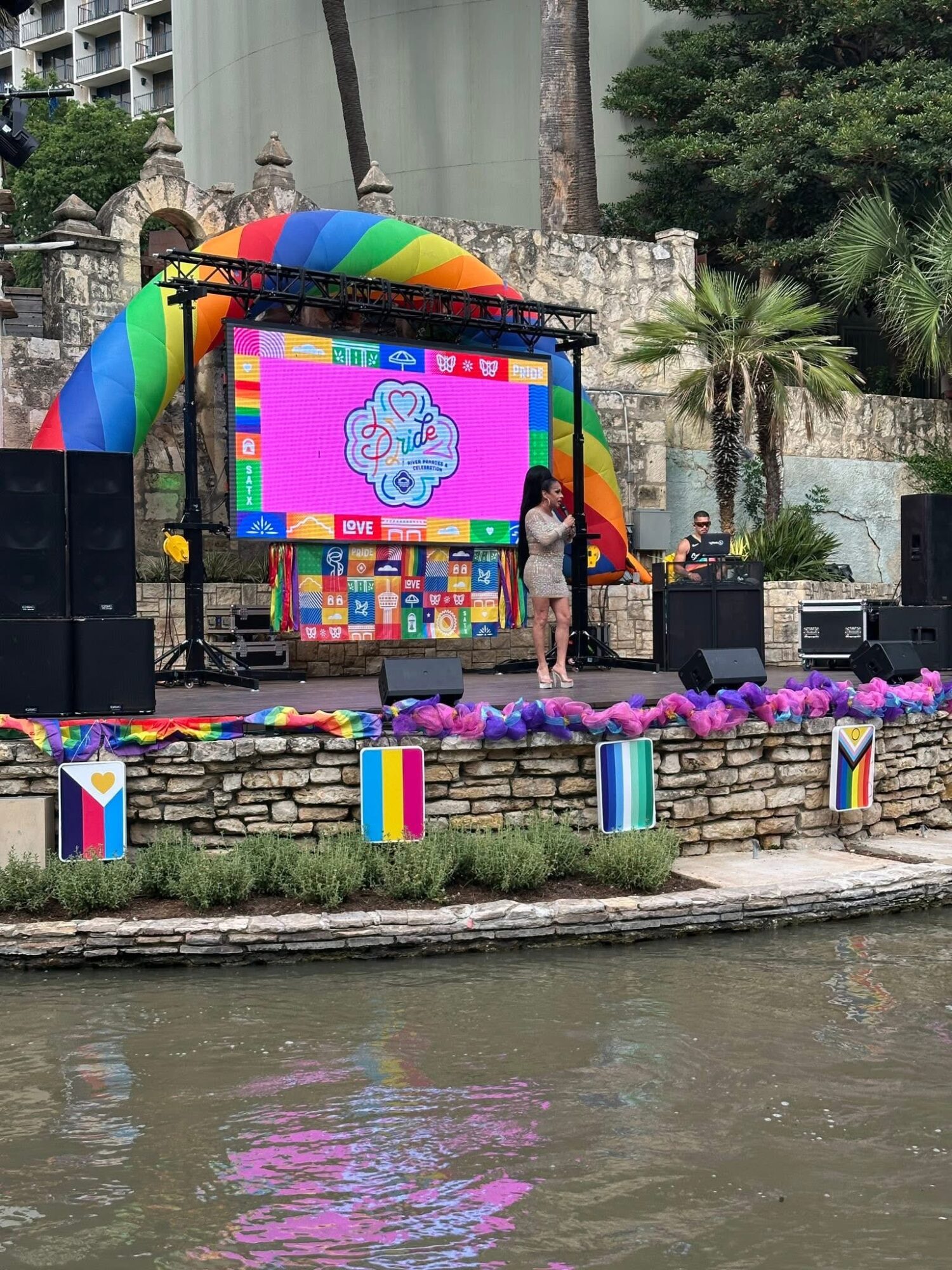 Stage with a large screen displaying a colorful cartoon character, decorated with rainbow and colorful banners, set outdoors near water.