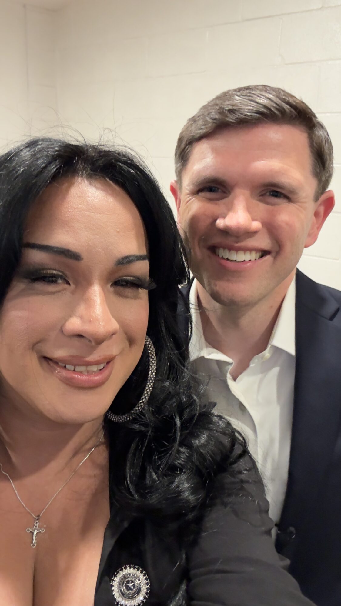 Two people smiling, one woman with dark hair and a man with light hair, in formal attire, indoors.