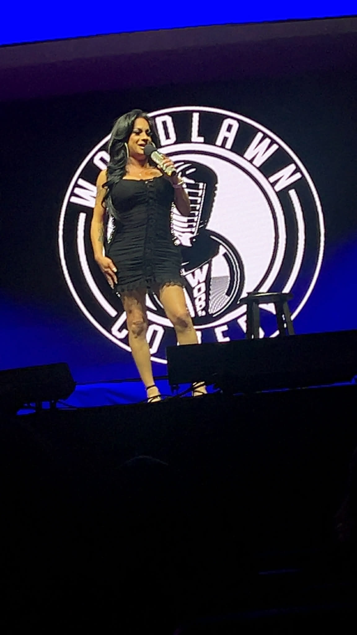 Woman standing on stage in front of a large circular logo with text, wearing a black dress and high heels.