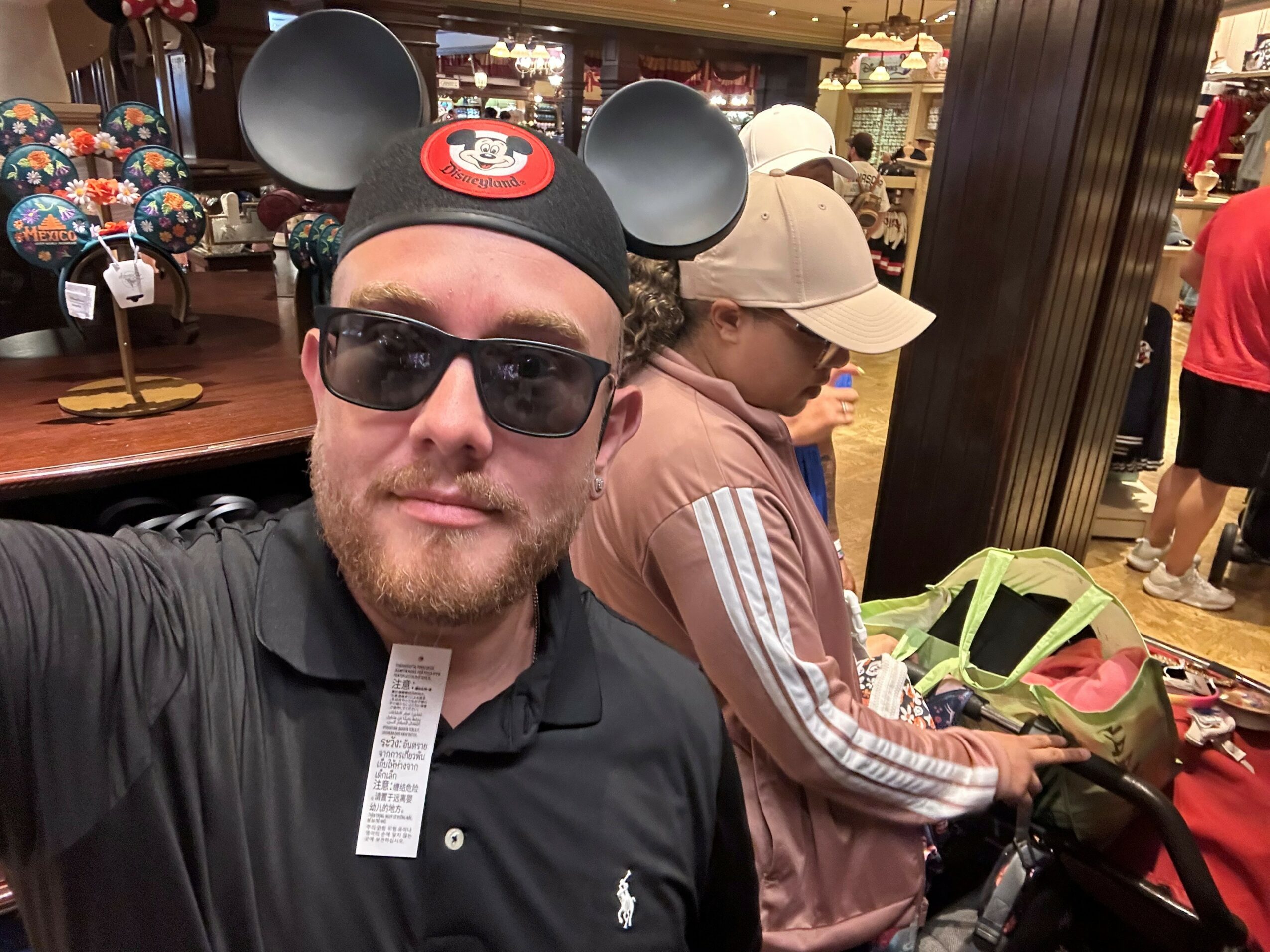 Man wearing Mickey Mouse ears, sunglasses, and a black shirt, taking a selfie in a store with people shopping in the background.