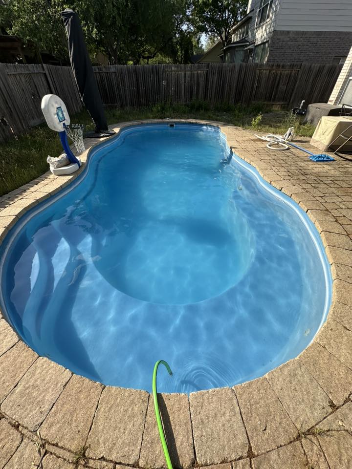 In-ground swimming pool with water, surrounded by a brick patio, fence, and outdoor equipment in a backyard.