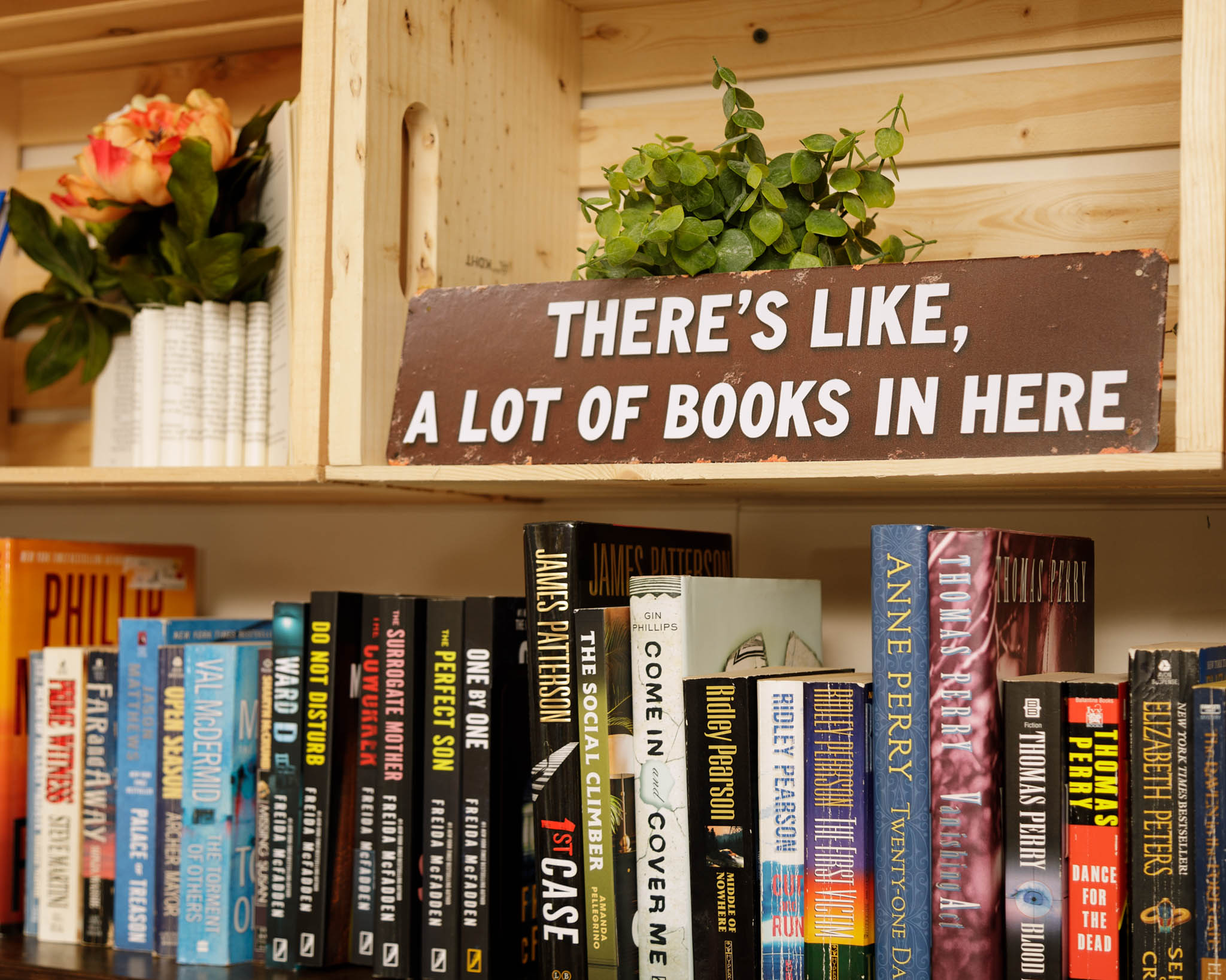 Bookshelves filled with books, a sign reading 'THERE'S LIKE, A LOT OF BOOKS IN HERE,' a potted plant, and a flower arrangement.