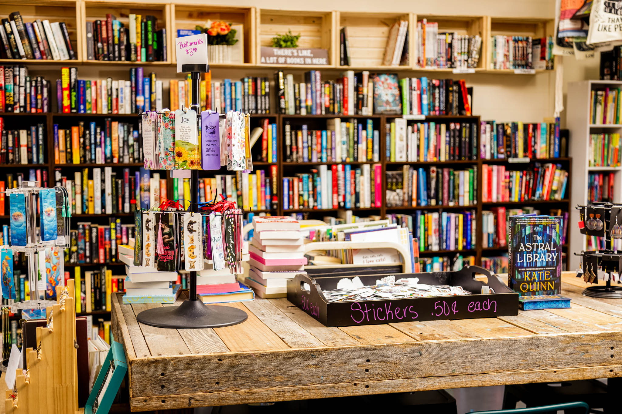Bookshelves filled with books behind a wooden table with stacks of books and a black tray, in a bookstore or library.