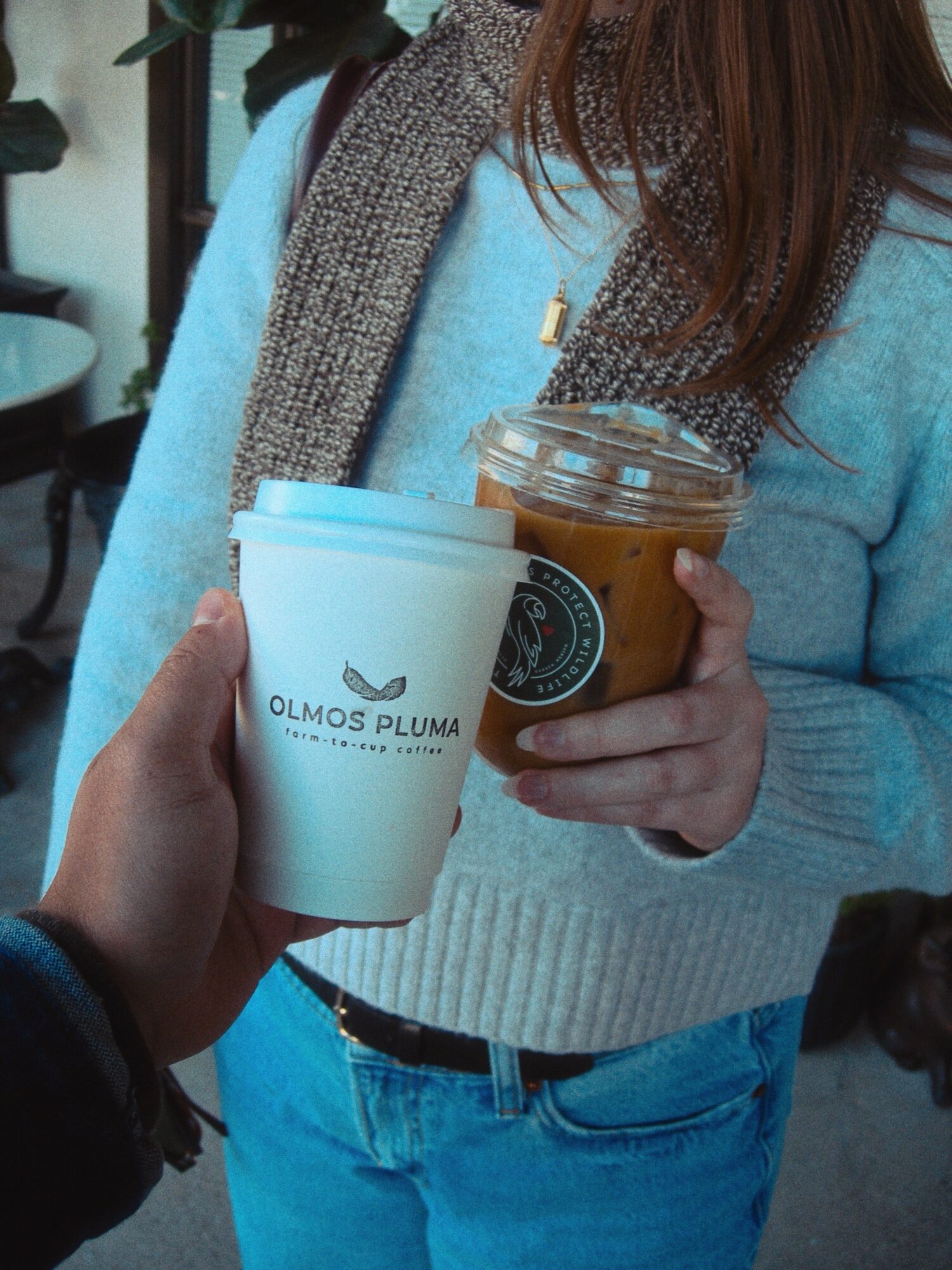 Person holding a plastic cup of iced coffee, wearing a blue sweater and gray scarf, with another hand holding a white paper cup.