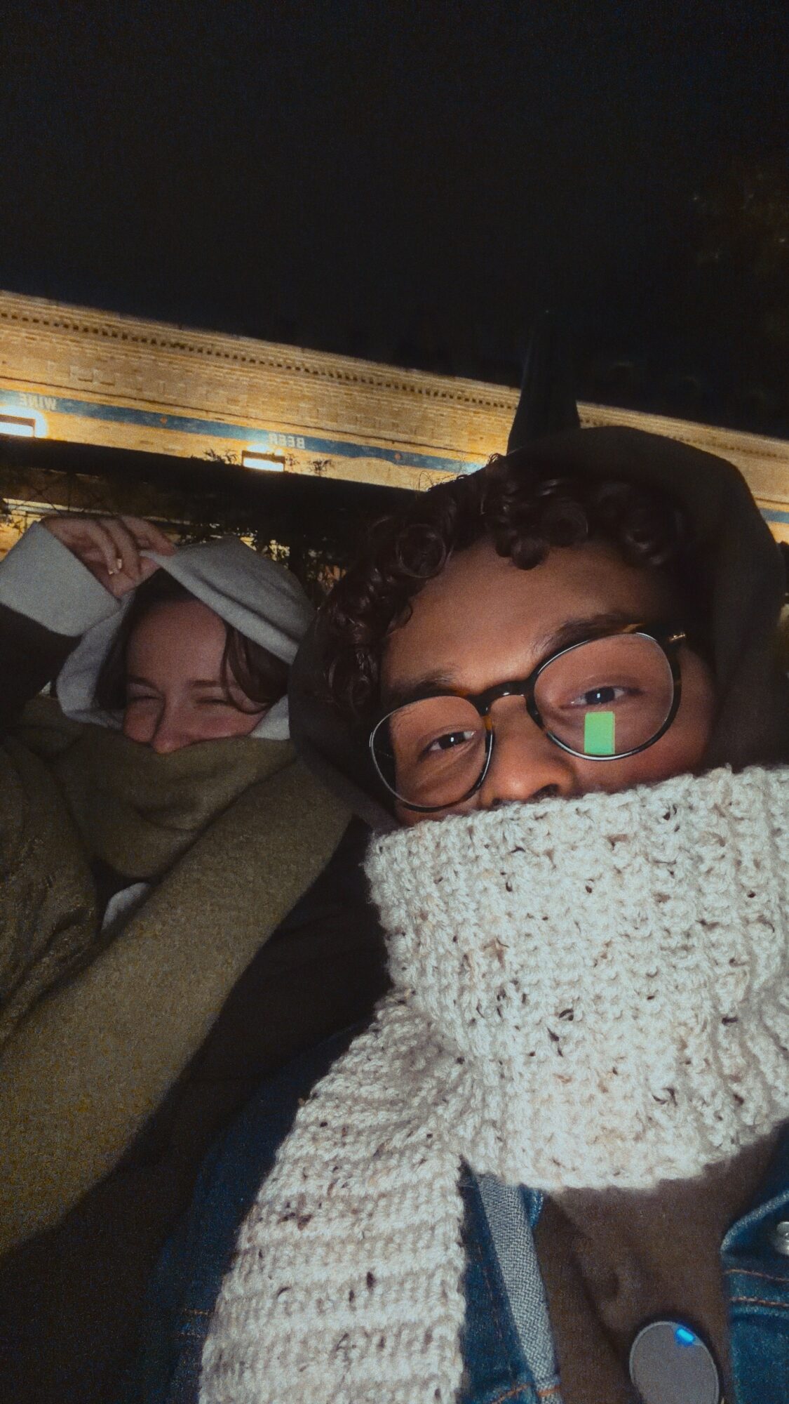 Two people bundled in warm clothing, sitting close together in a dark setting, with one person wearing glasses and a scarf.