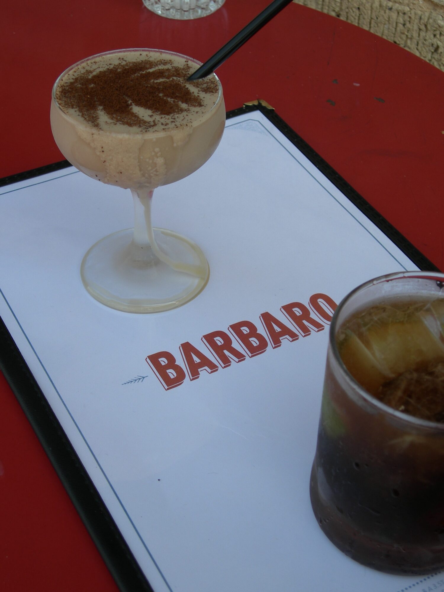 Two drinks on a menu with the word 'BARBARO' in red, one with a straw and frothy top, the other dark with ice.