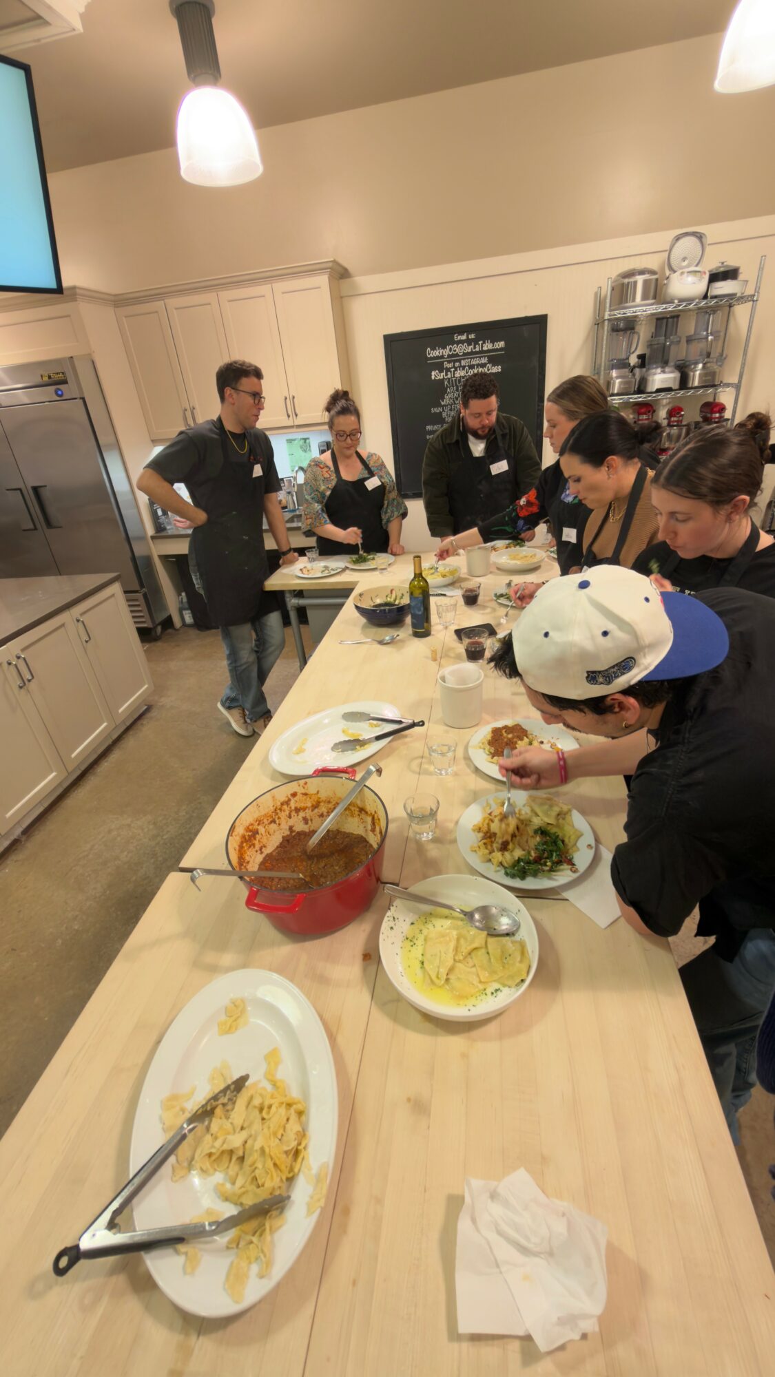 Group of people gathered around a kitchen island with food, some standing and some sitting, in a modern kitchen.