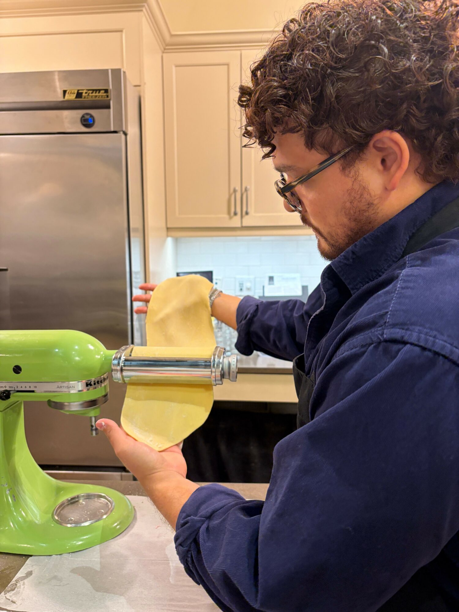 Person with curly hair and glasses uses a green kitchen appliance, possibly a juicer, on a countertop in a kitchen.