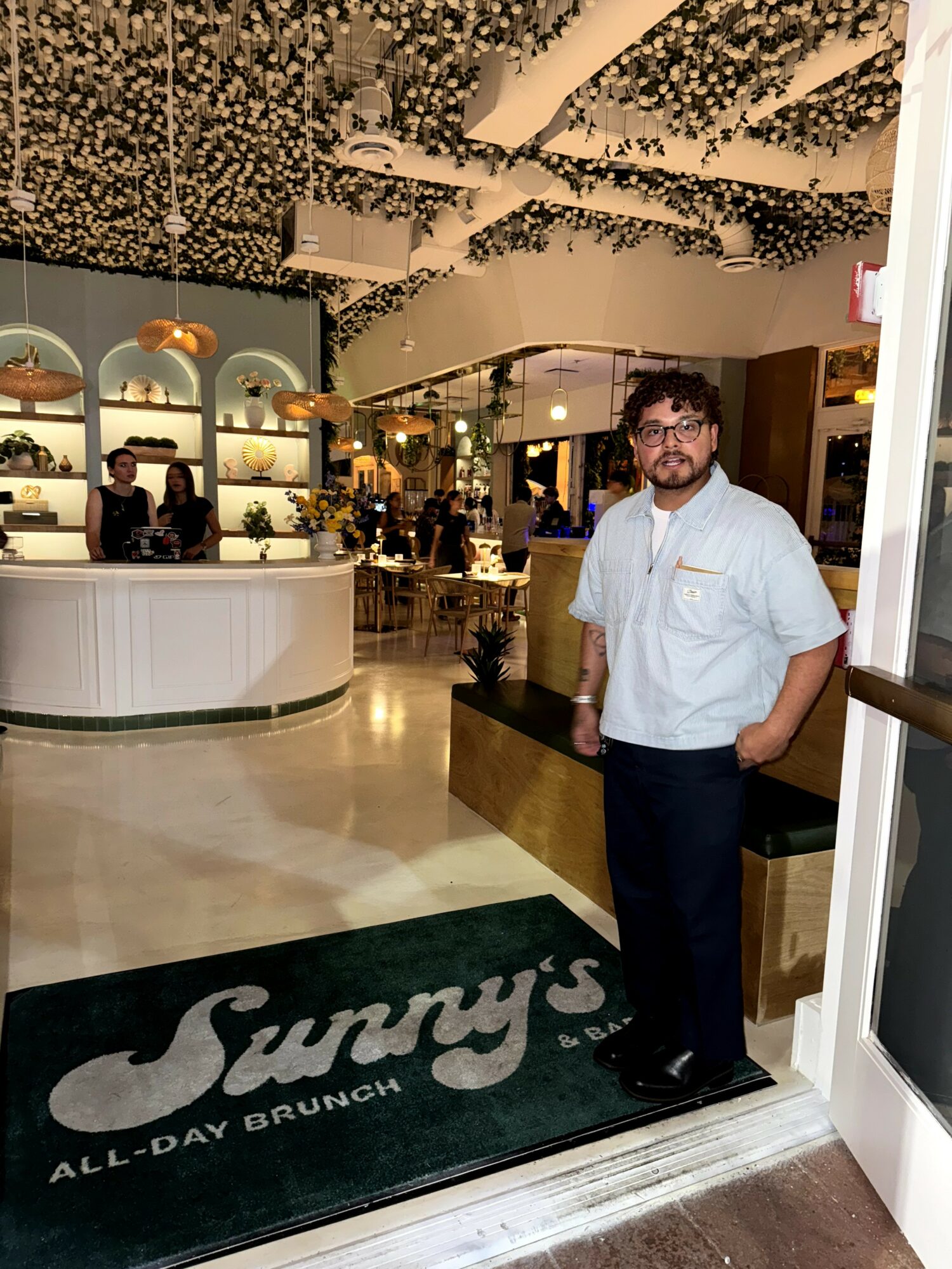 Man with glasses and beard in white shirt standing inside a restaurant or cafe, with a counter and decorative ceiling, smiling.