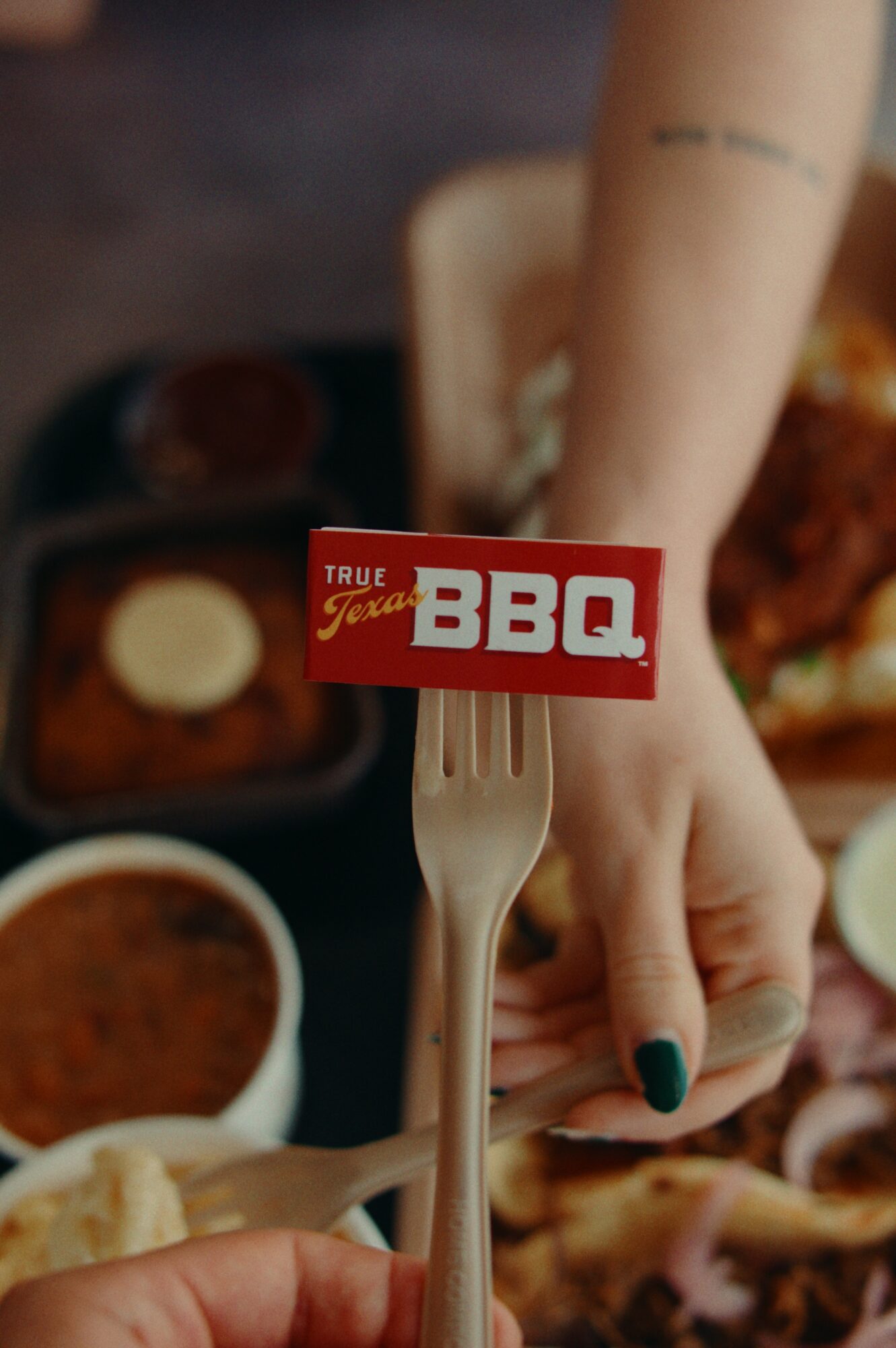 Person holding a fork with a red BBQ sign, surrounded by food dishes, arm with tattoo visible.