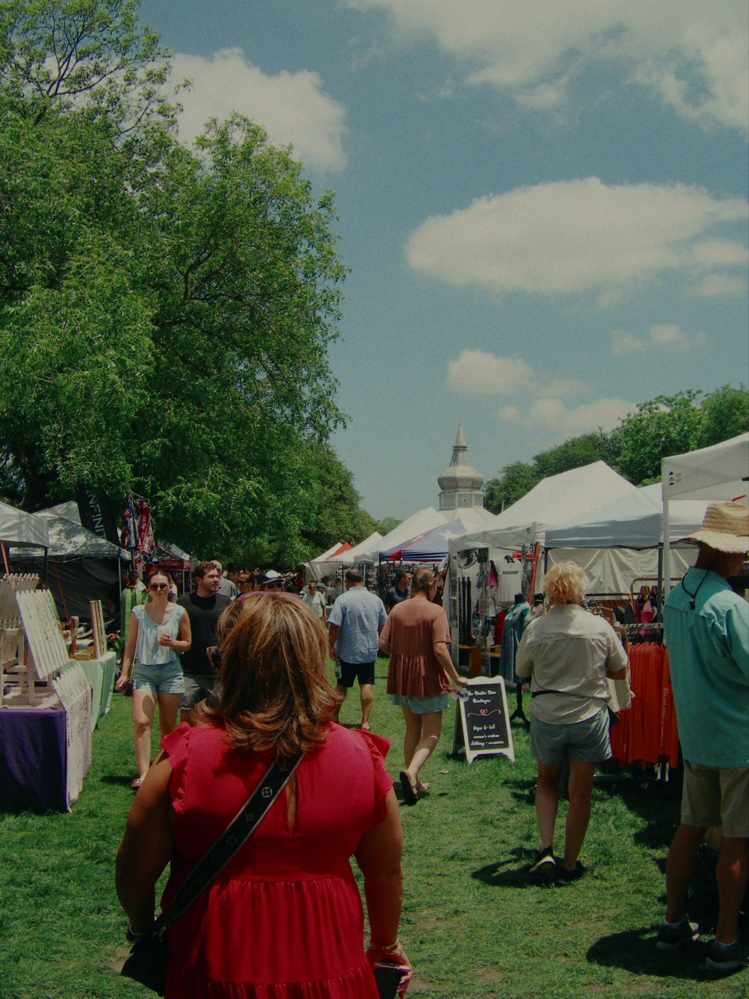 People walking outdoors at a market or fair with tents and trees, blue sky with clouds in background.