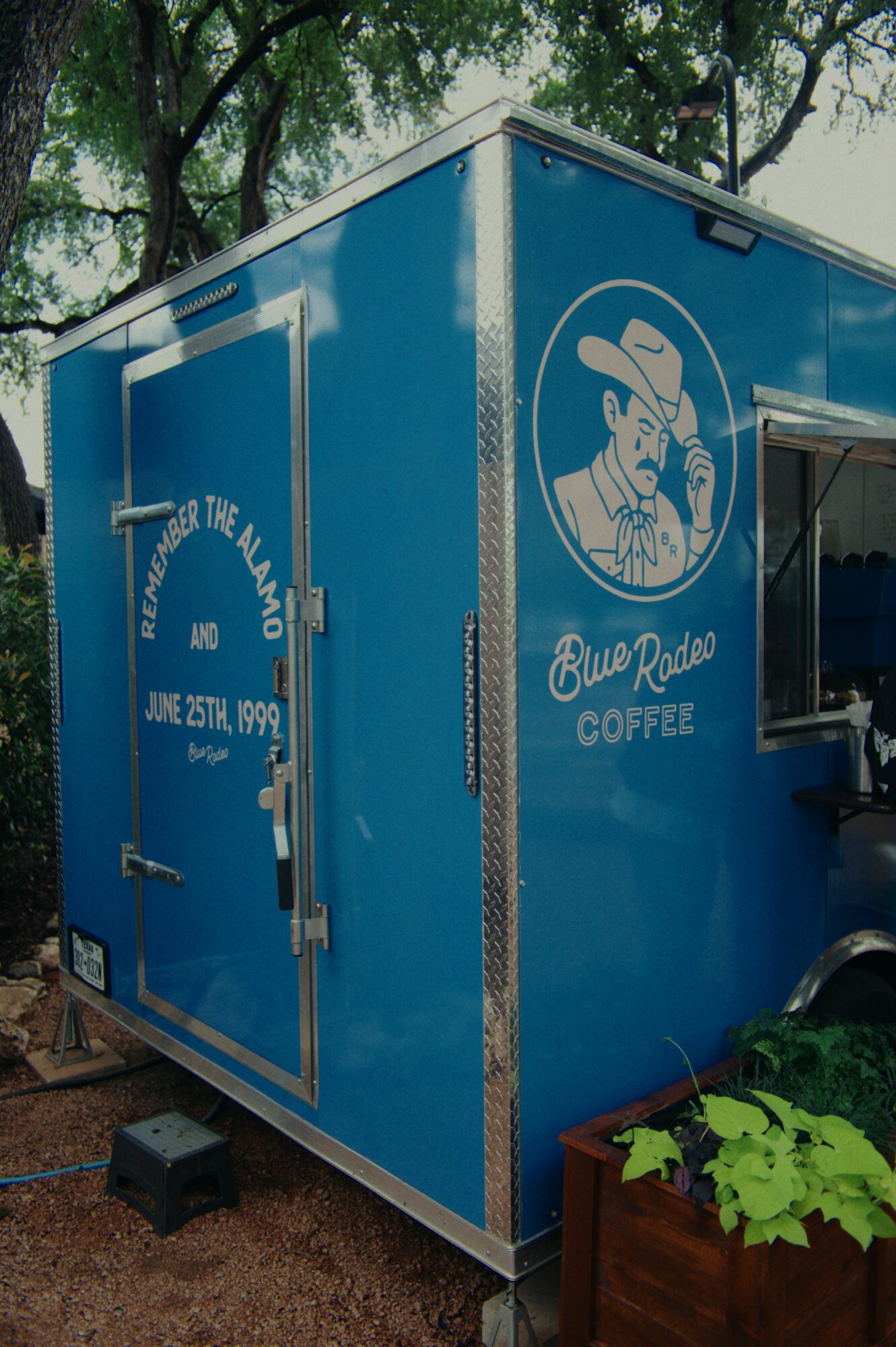 Blue mobile coffee trailer with logo of a man in a hat holding a coffee cup, outdoors with trees in background.