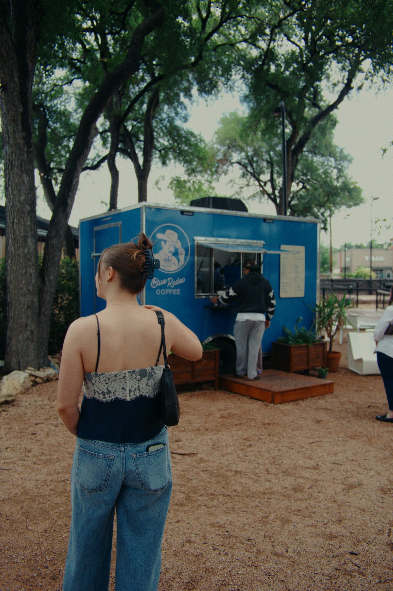 Person with short hair, wearing a black and white top and blue jeans, stands outdoors near a blue booth with trees in background.
