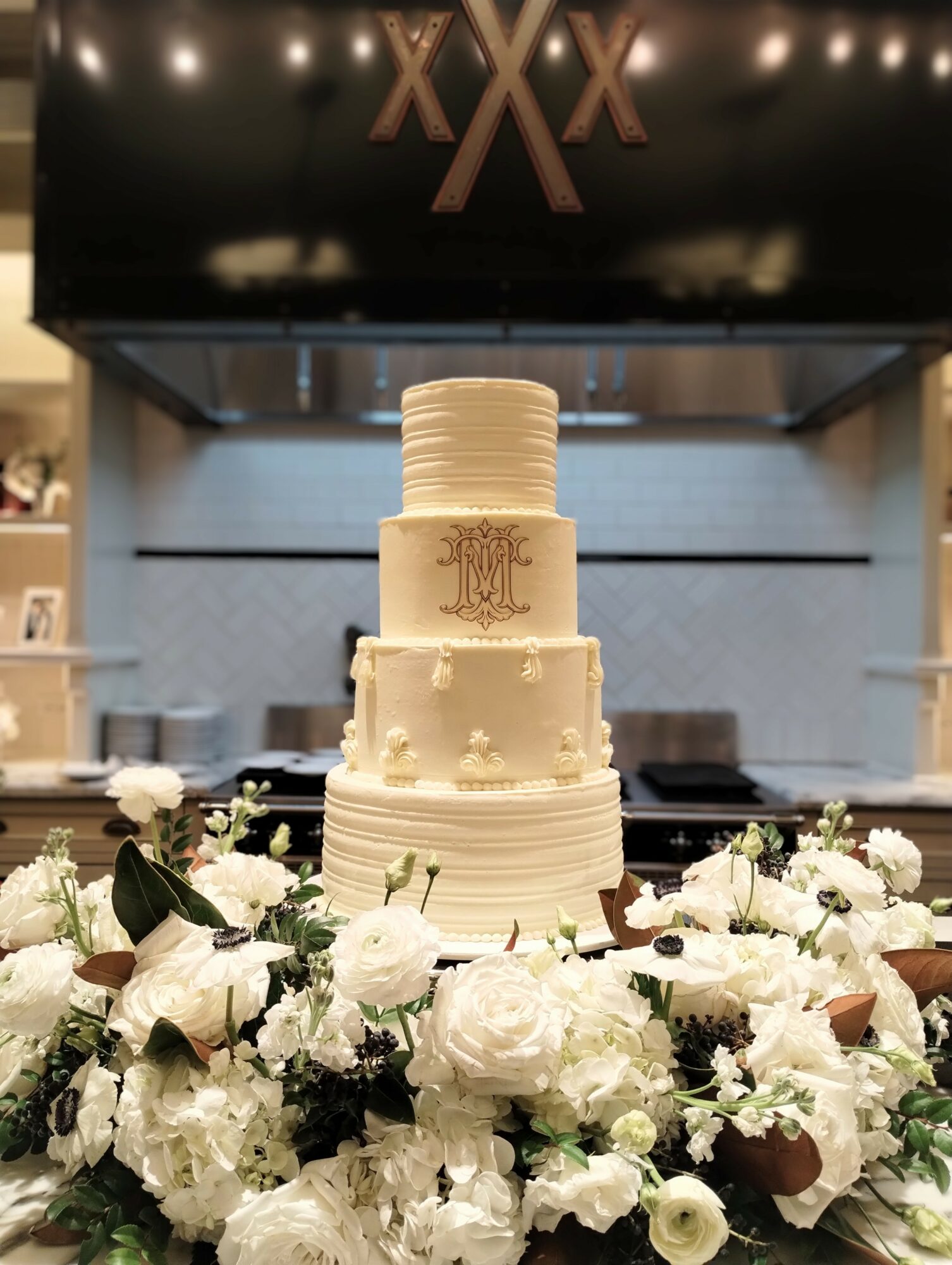 Three-tiered white wedding cake with gold monogram, surrounded by white flowers, in a decorated venue.