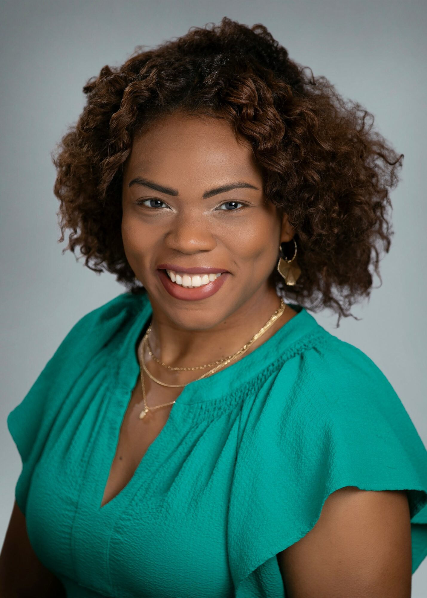 Smiling woman with curly hair wearing a teal blouse and gold jewelry, against a neutral background.