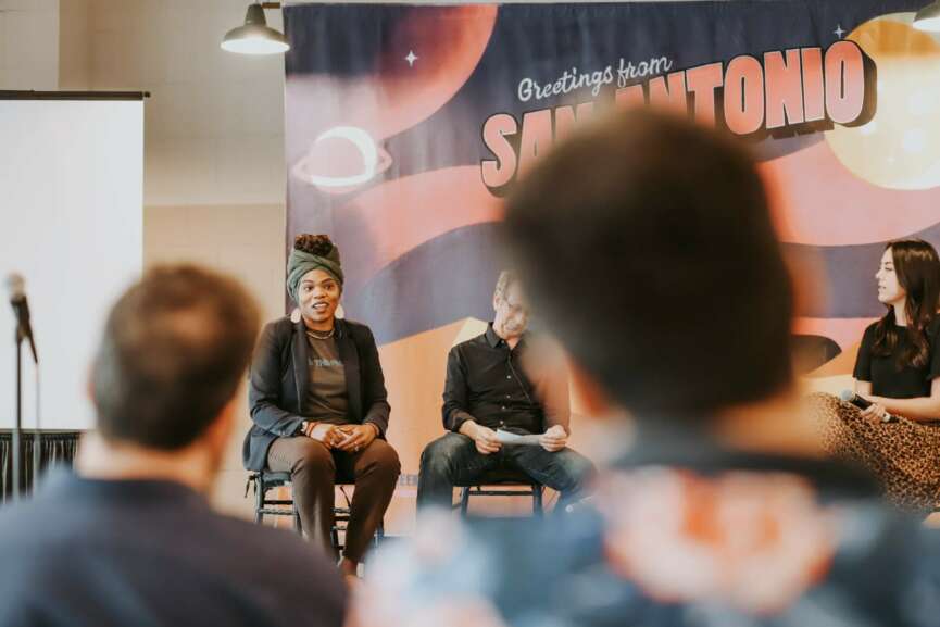 Three women sit on a stage with a space-themed backdrop, speaking to an audience. Audience members' heads are visible in foreground.