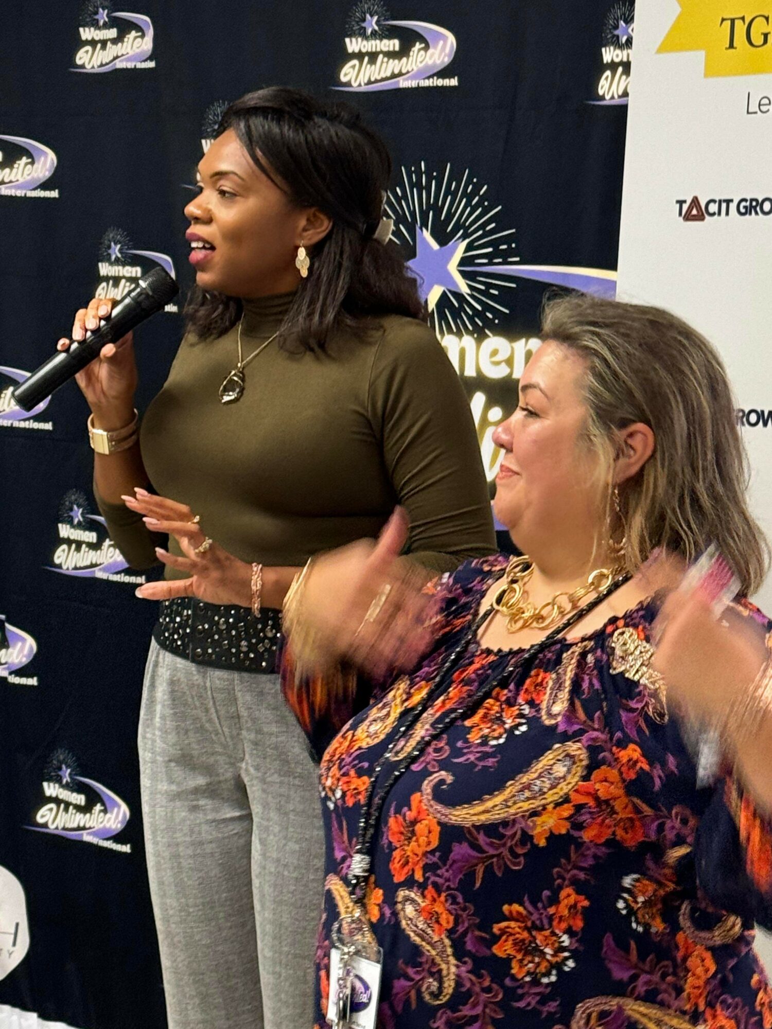 Two women speaking at an event, one holding a microphone, with a dark backdrop and event banners behind them.