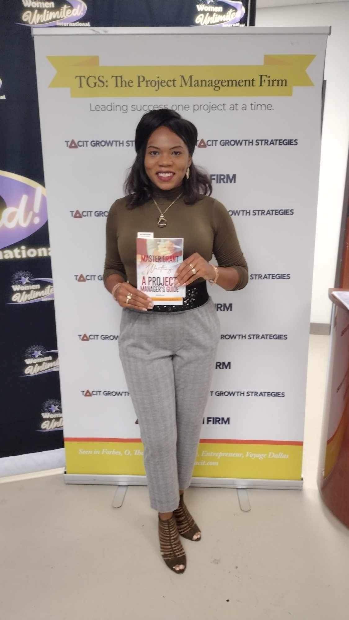 Woman holding a book in front of a banner, smiling, wearing a brown top and gray pants, in an indoor setting.
