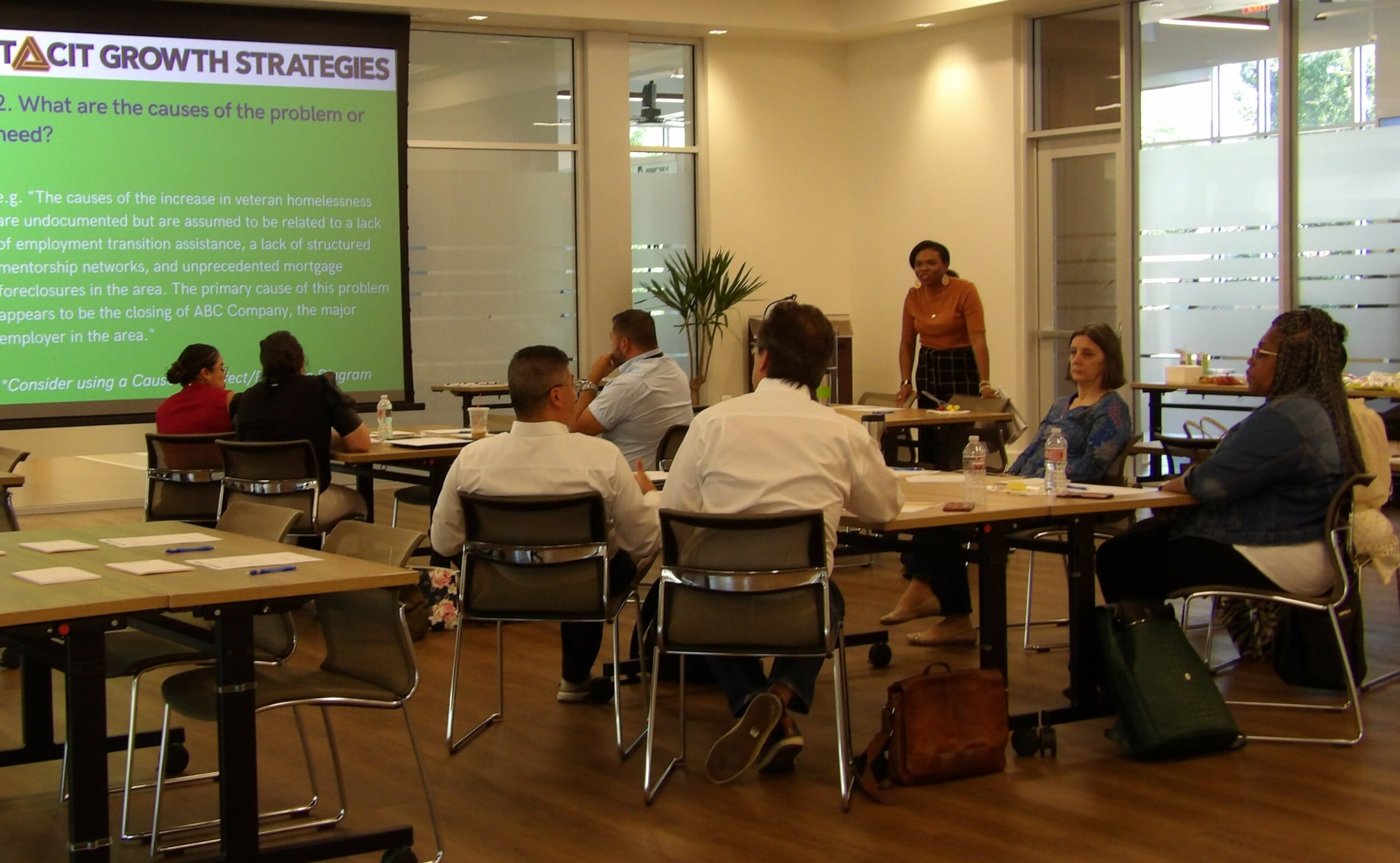 People seated around tables in a conference room, a presenter standing near a large screen displaying a presentation, windows in the background.