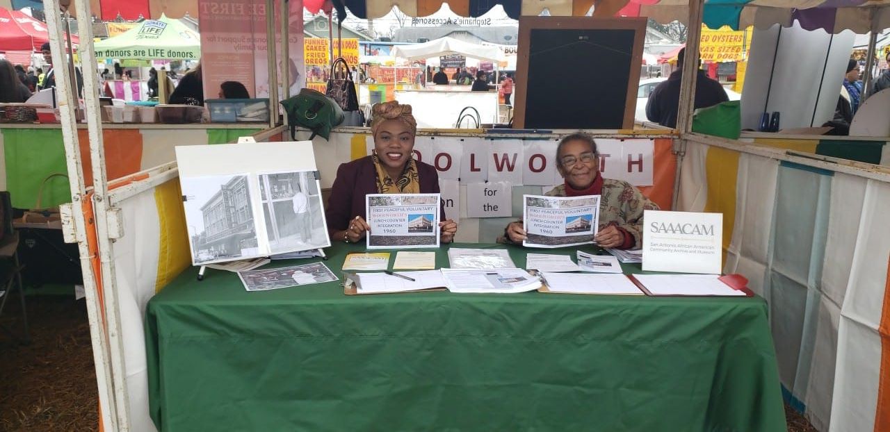 Two women sit behind a table at an outdoor event, with newspapers and informational materials displayed.