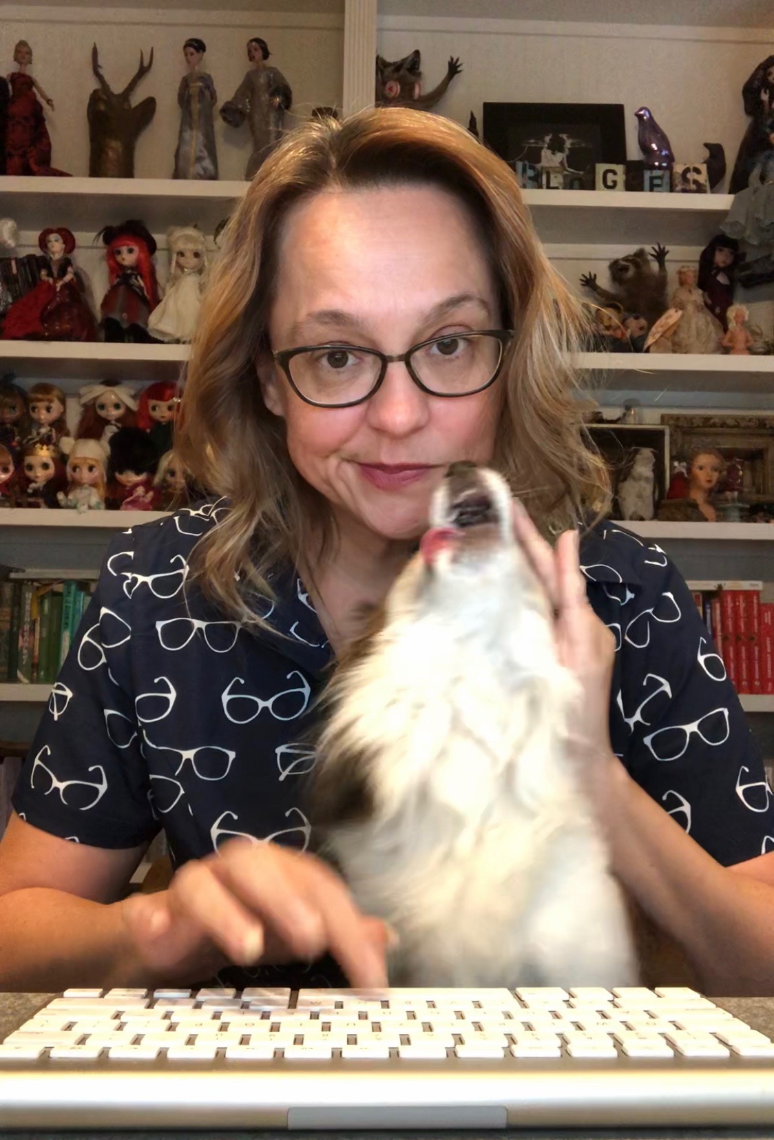 Woman with glasses and wavy hair sitting at a desk with a white fluffy dog, shelves with dolls and books in background.